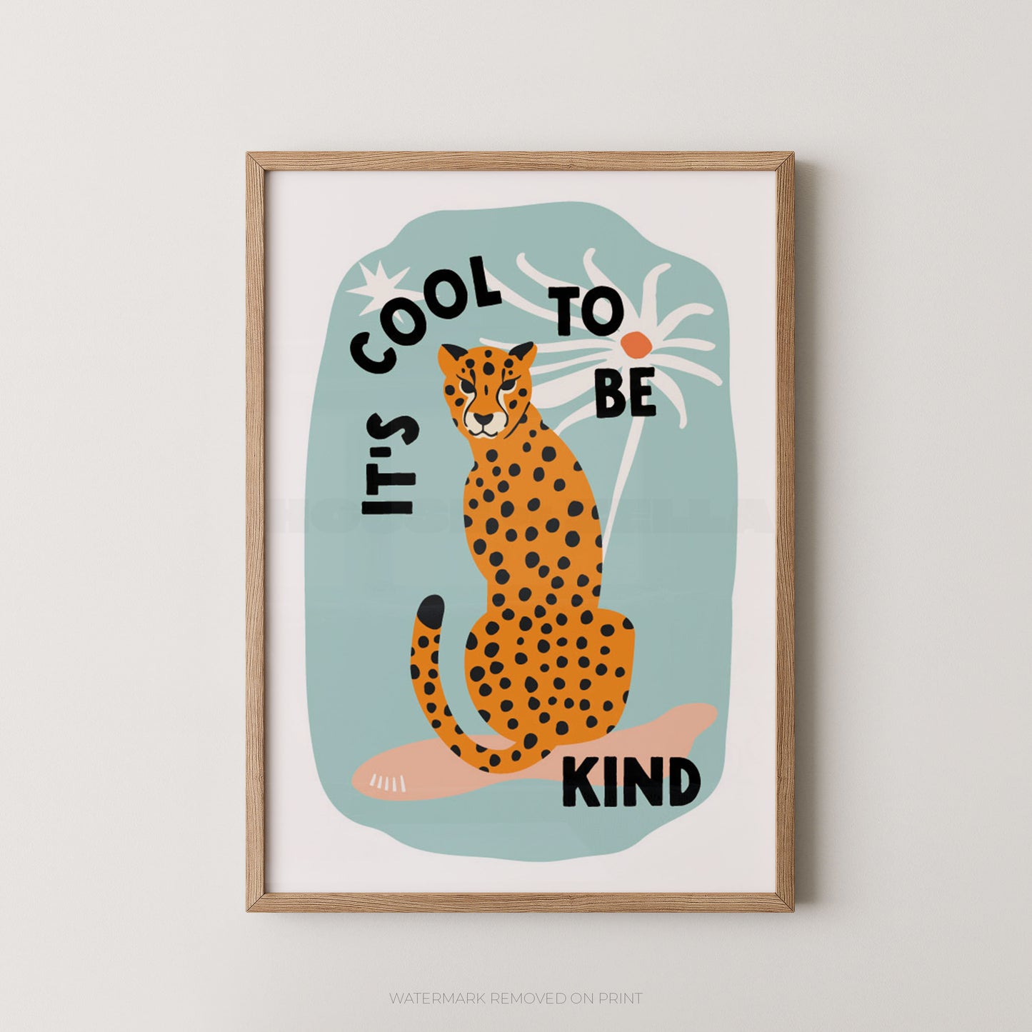A framed art print featuring an illustrated orange-and-black spotted cat sitting on a pale blue shape with the text 'IT'S COOL TO BE KIND' in black lettering, shown against a light neutral wall.