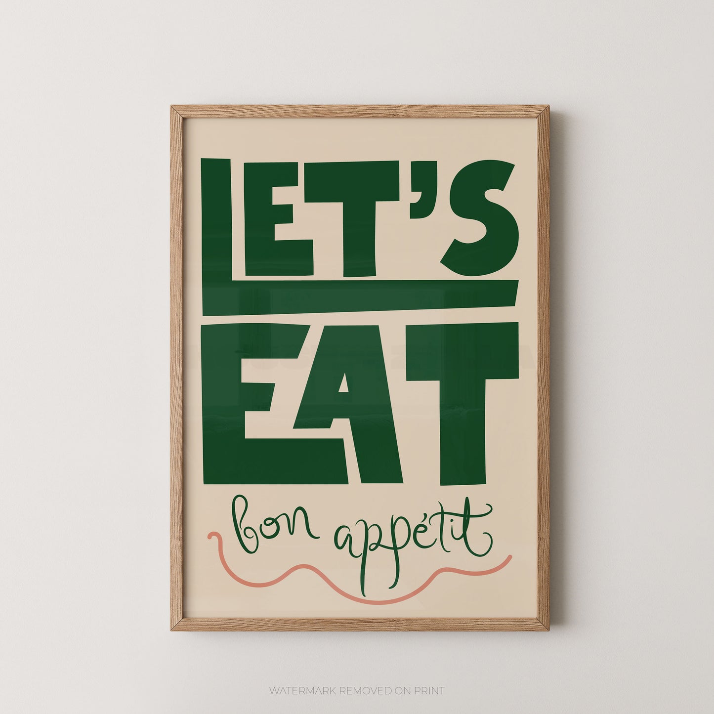A framed typographic wall art print with the words “LET’S EAT” in large dark green letters on a warm cream background, with “bon appetit” in smaller green script and a coral line drawing below. The print is shown in a light natural wood frame against a light gray wall.