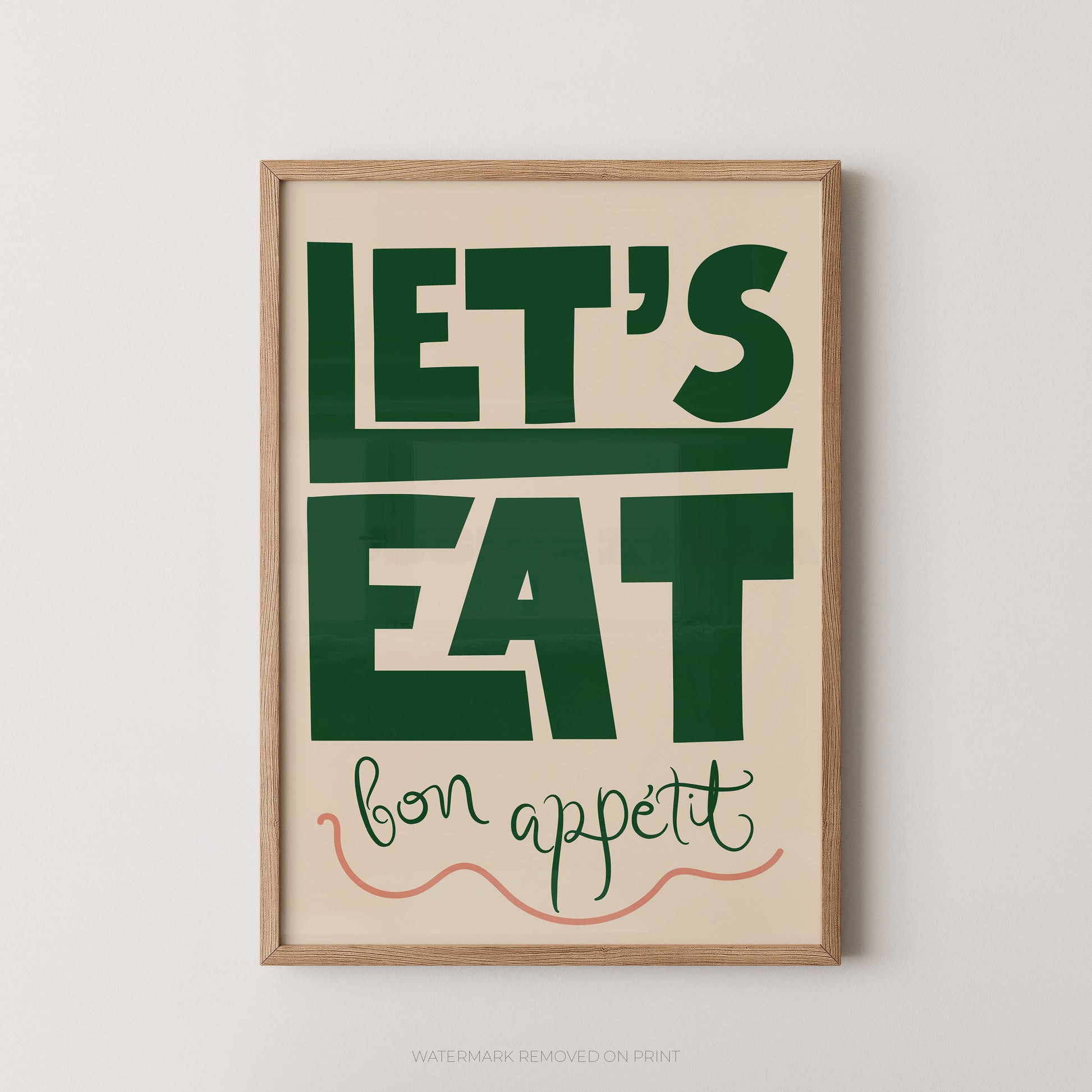 A framed typographic wall art print with the words “LET’S EAT” in large dark green letters on a warm cream background, with “bon appetit” in smaller green script and a coral line drawing below. The print is shown in a light natural wood frame against a light gray wall.