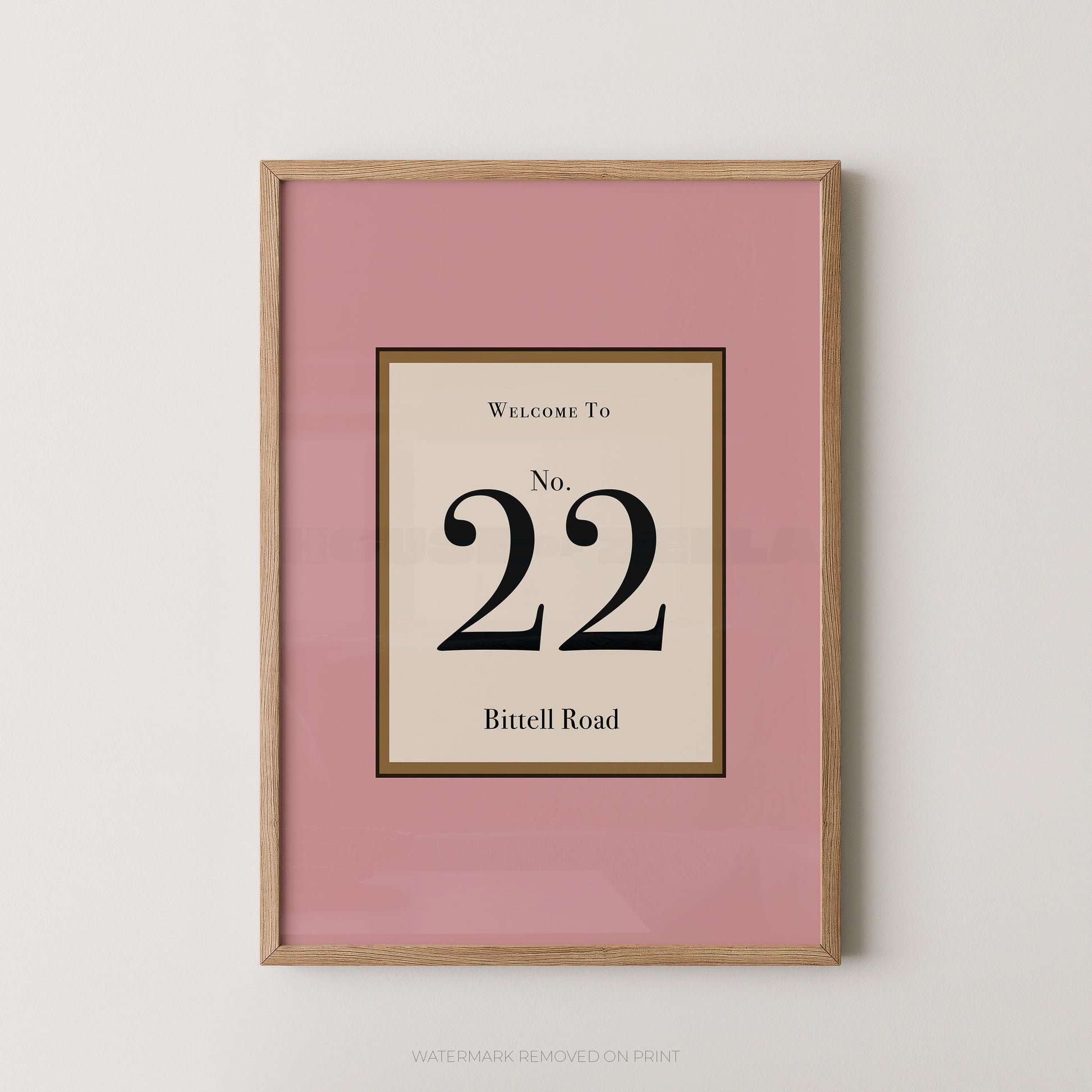 Framed wall art print with a pink border and a central cream panel reading 'Welcome to No. 22 Bittell Road' in black text, shown in a light wood frame against a light gray wall.