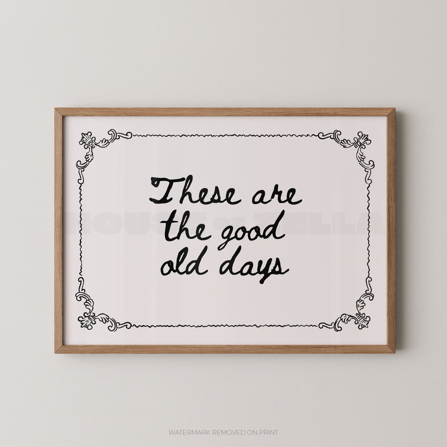 Framed wall art print with the phrase “These are the good old days” in black script inside an ornate black border, set in a light natural wood frame against a light gray wall.
