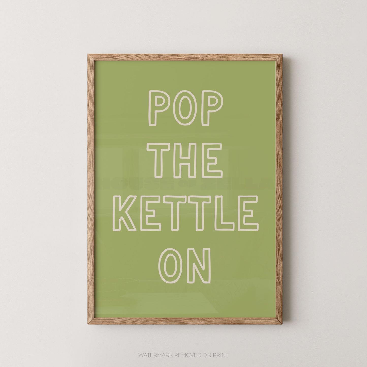 A framed typographic wall art print with the phrase “POP THE KETTLE ON” in white outlined letters on a muted green background, shown hanging against a light neutral wall.