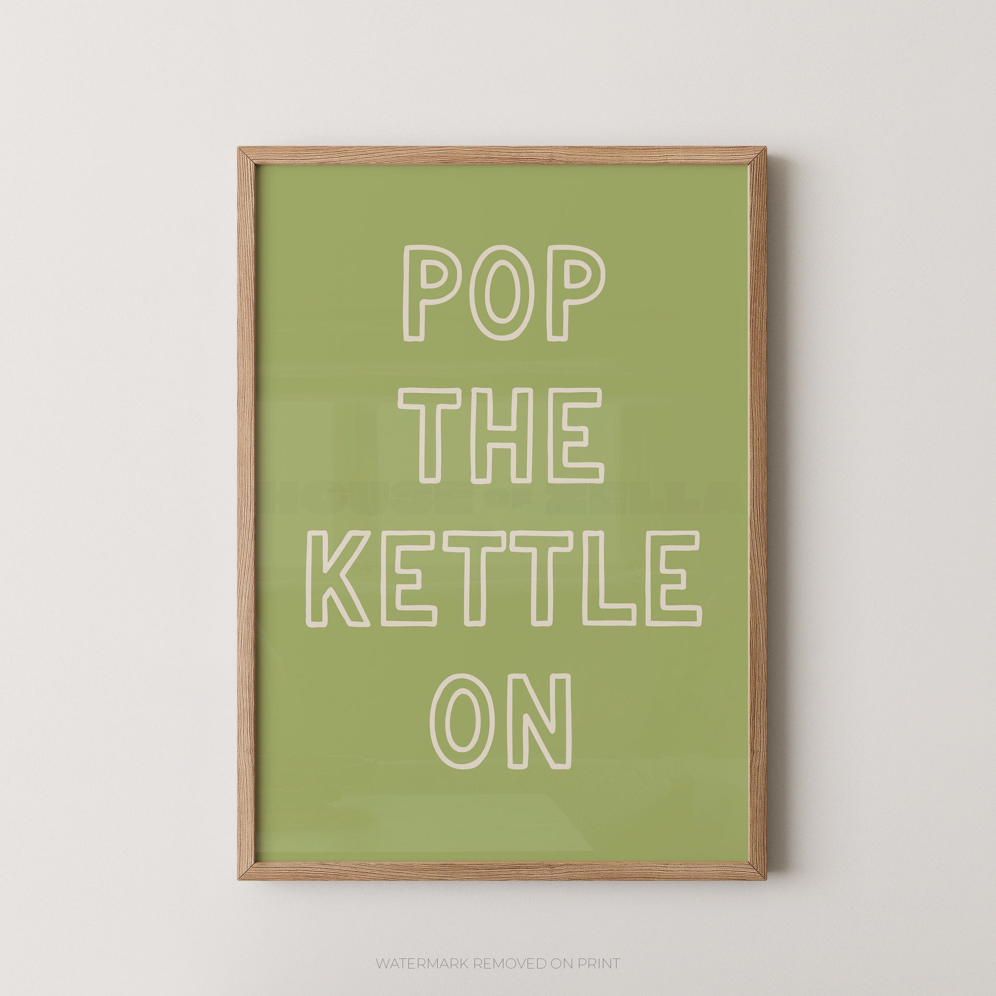 A framed typographic wall art print with the phrase “POP THE KETTLE ON” in white outlined letters on a muted green background, shown hanging against a light neutral wall.