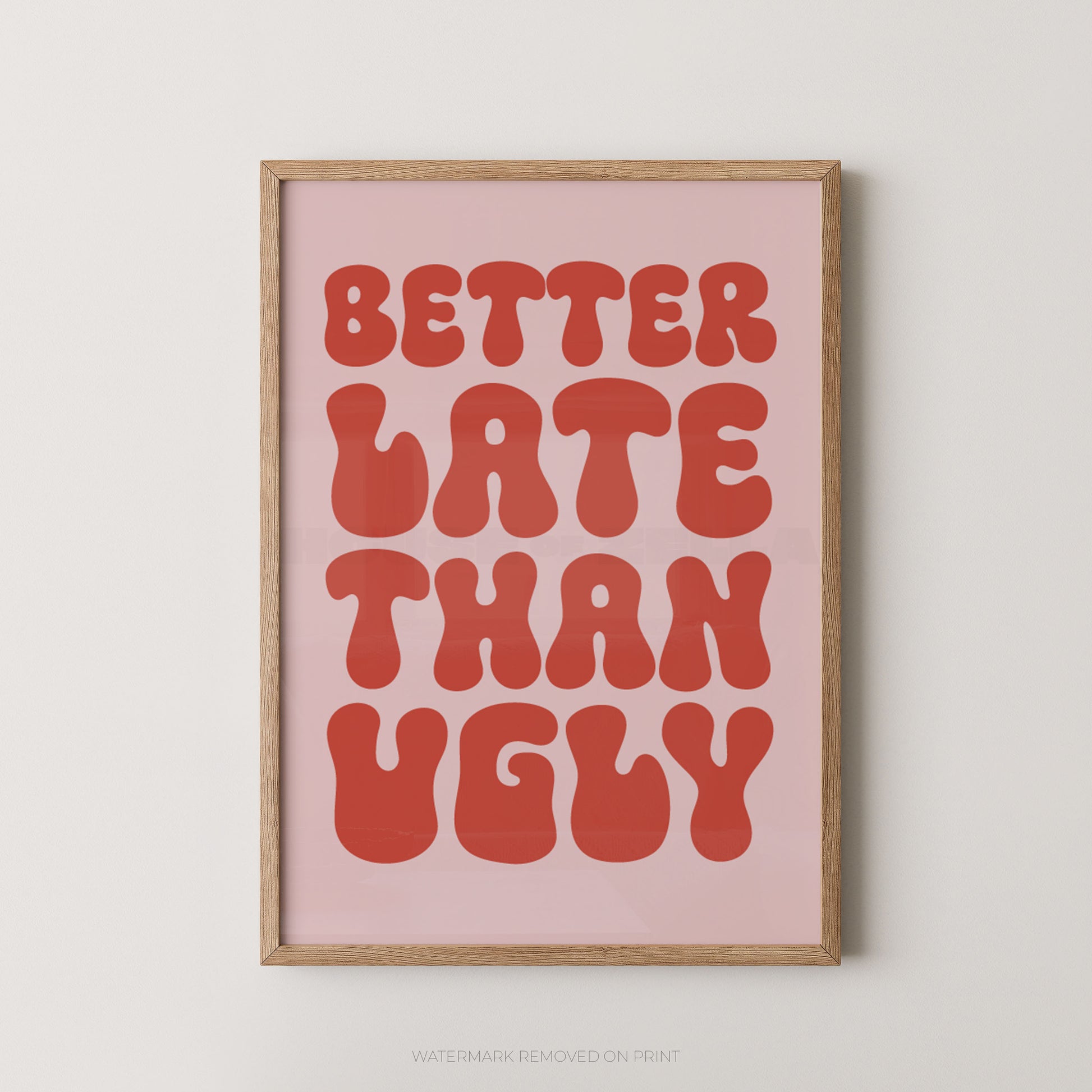 A framed typographic wall art print with the phrase “BETTER LATE THAN UGLY” in large coral-red retro letters on a pale pink background, shown in a light natural wood frame against a light gray wall.