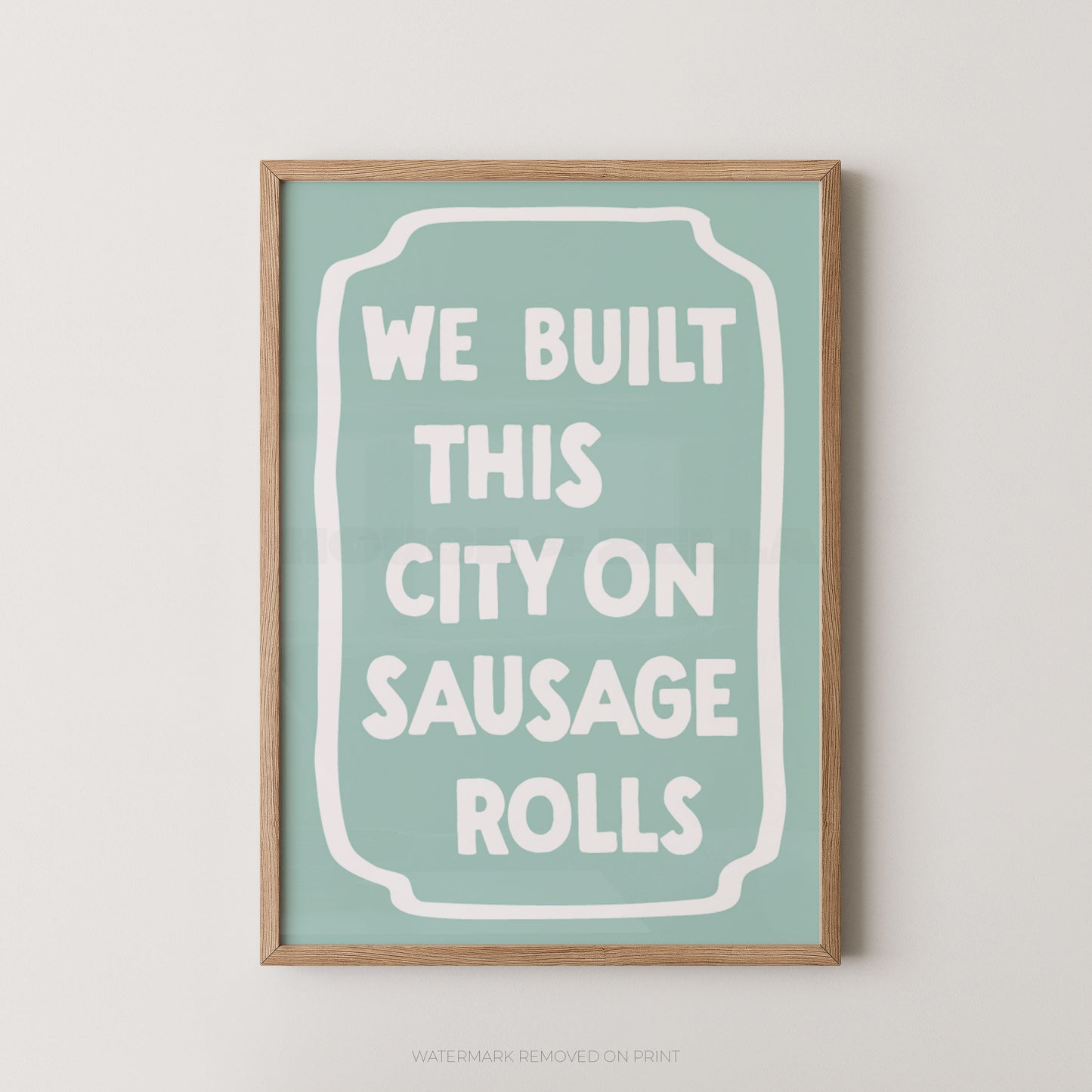 A framed typographic wall art print with a light wood frame, a pale mint background, and white text reading “WE BUILT THIS CITY ON SAUSAGE ROLLS.”.