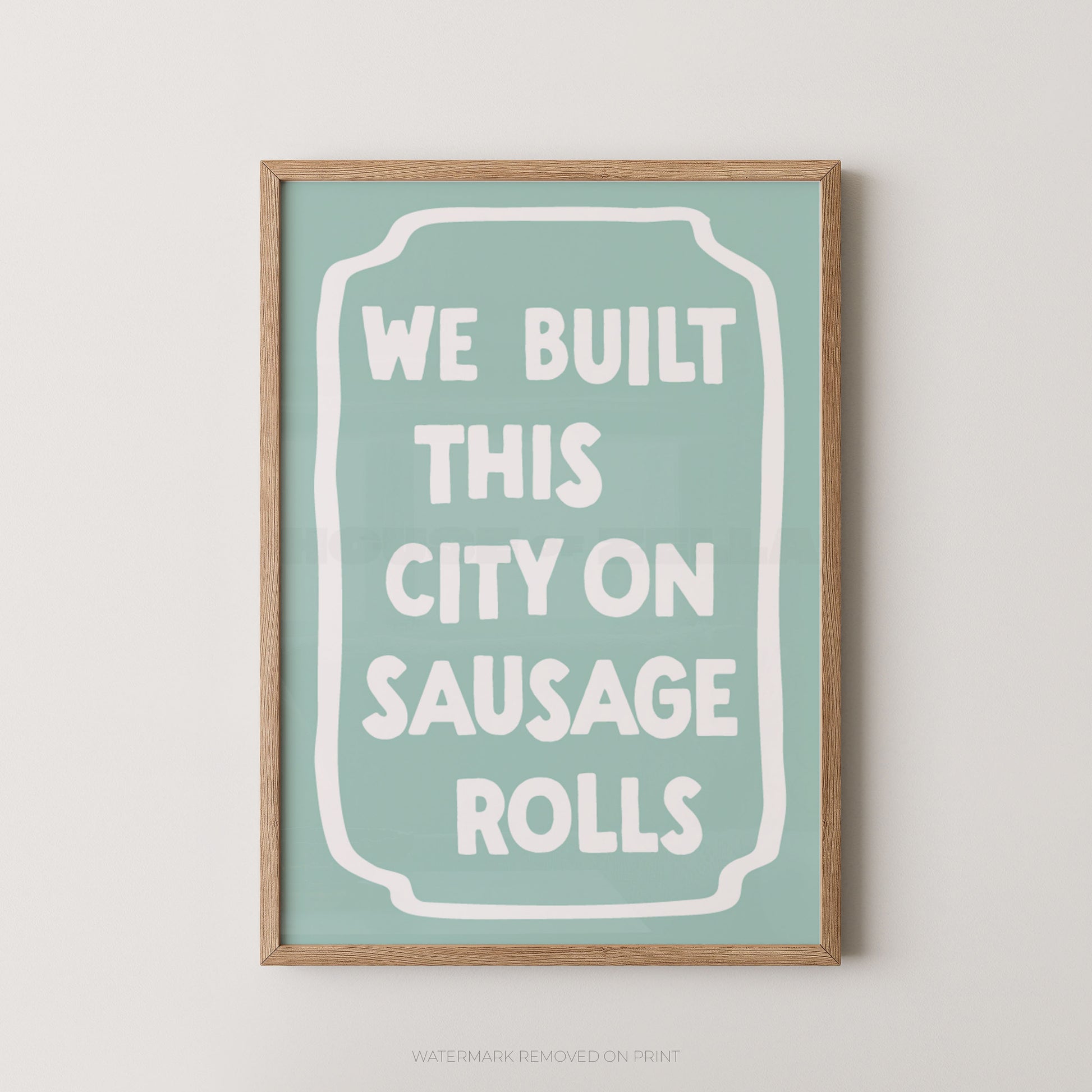 A framed typographic wall art print with a light wood frame, a pale mint background, and white text reading “WE BUILT THIS CITY ON SAUSAGE ROLLS.”.