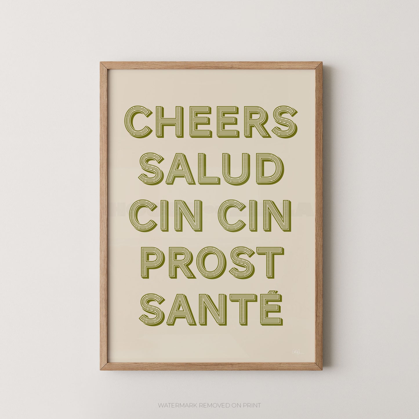 Framed typographic wall art with the phrase “CHEERS SALUD CIN CIN PROST SANTÉ” in olive-green raised letters on a warm cream background, shown in a light natural wood frame against a light gray wall.