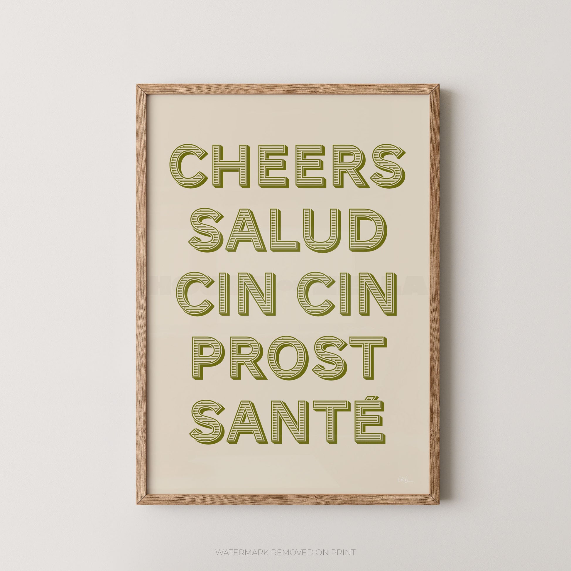 Framed typographic wall art with the phrase “CHEERS SALUD CIN CIN PROST SANTÉ” in olive-green raised letters on a warm cream background, shown in a light natural wood frame against a light gray wall.