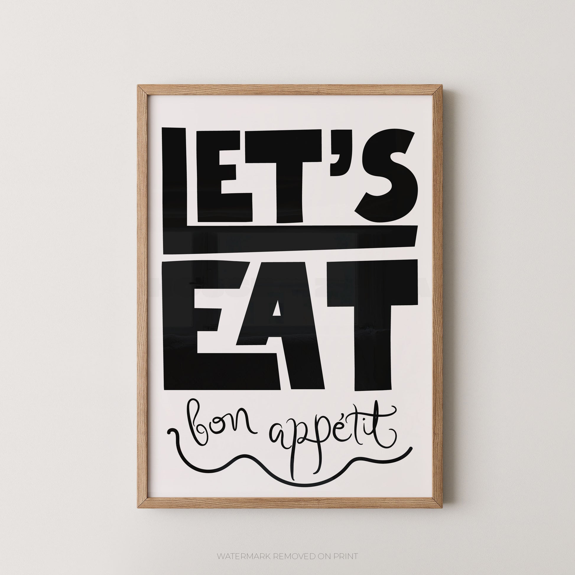 Framed typographic wall art with the words “LET’S EAT” in large black block letters and “bon appetit” in black script on a white background, shown in a light wood frame against a light gray wall.