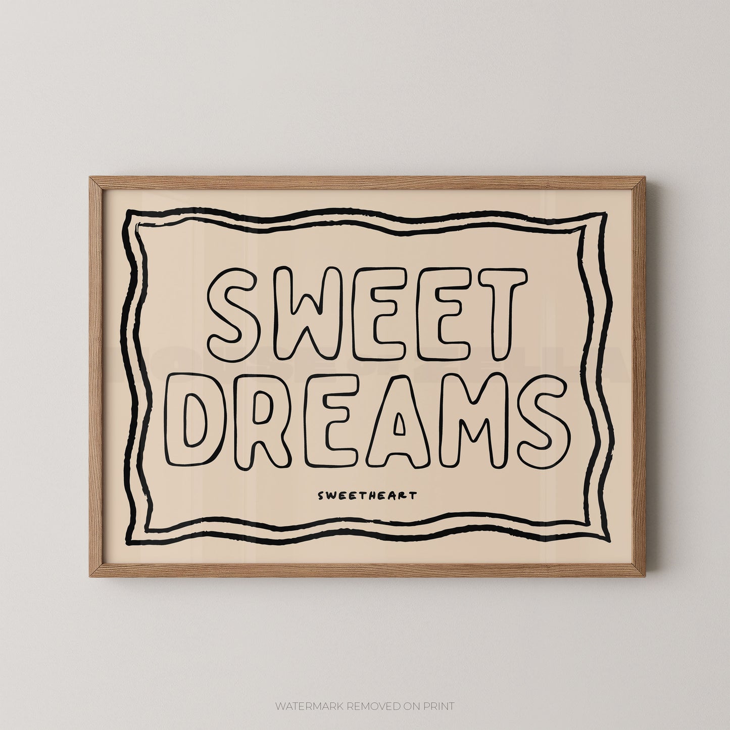 A framed typographic wall art print reading “SWEET DREAMS” with a small “SWEETHEART” mark below, shown hanging on a light gray wall. The print has a warm cream background with black lettering and border lines, and a light natural wood frame.