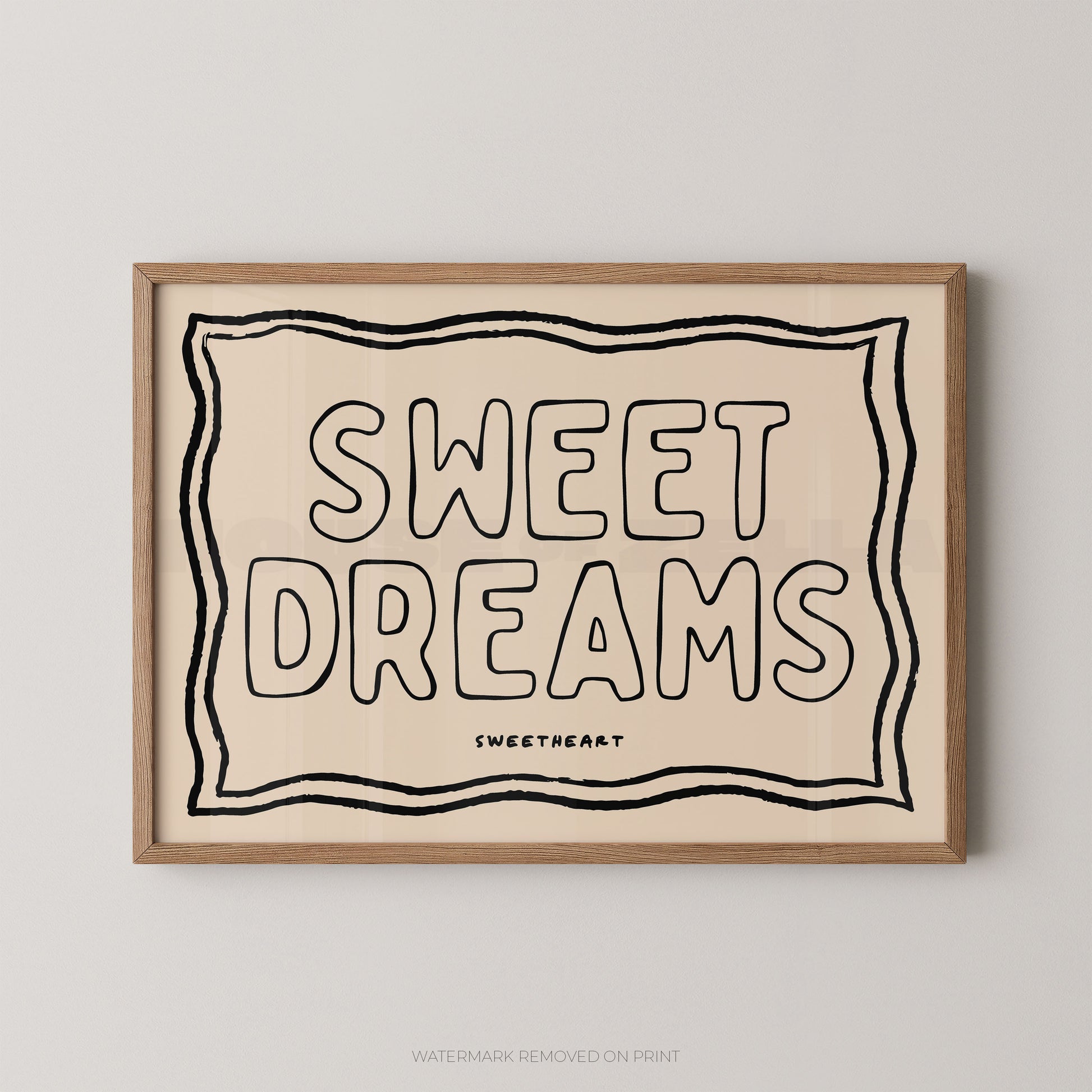 A framed typographic wall art print reading “SWEET DREAMS” with a small “SWEETHEART” mark below, shown hanging on a light gray wall. The print has a warm cream background with black lettering and border lines, and a light natural wood frame.