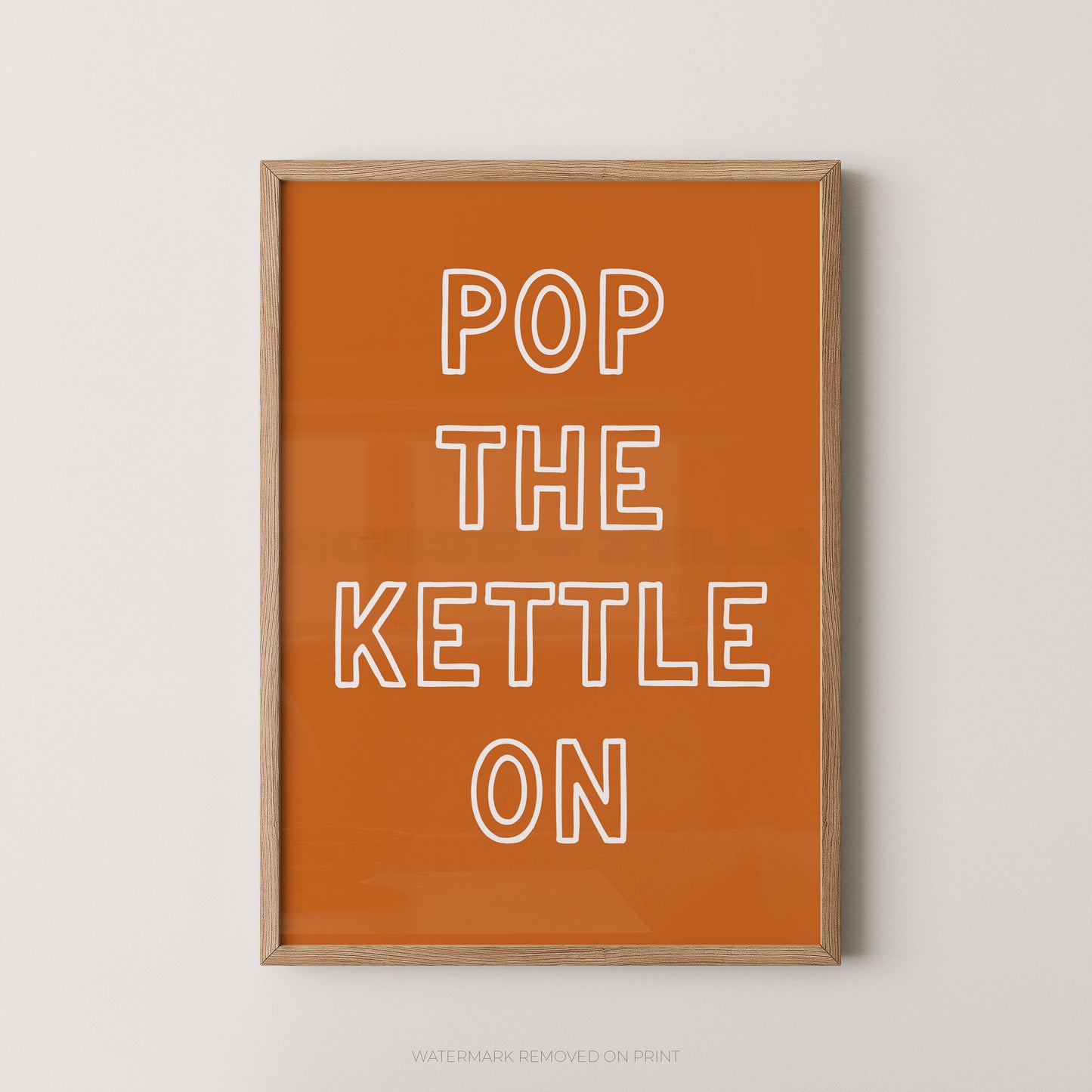 Framed wall art print with the phrase 'POP THE KETTLE ON' in white outlined letters on a warm orange background, shown hanging against a light neutral wall.