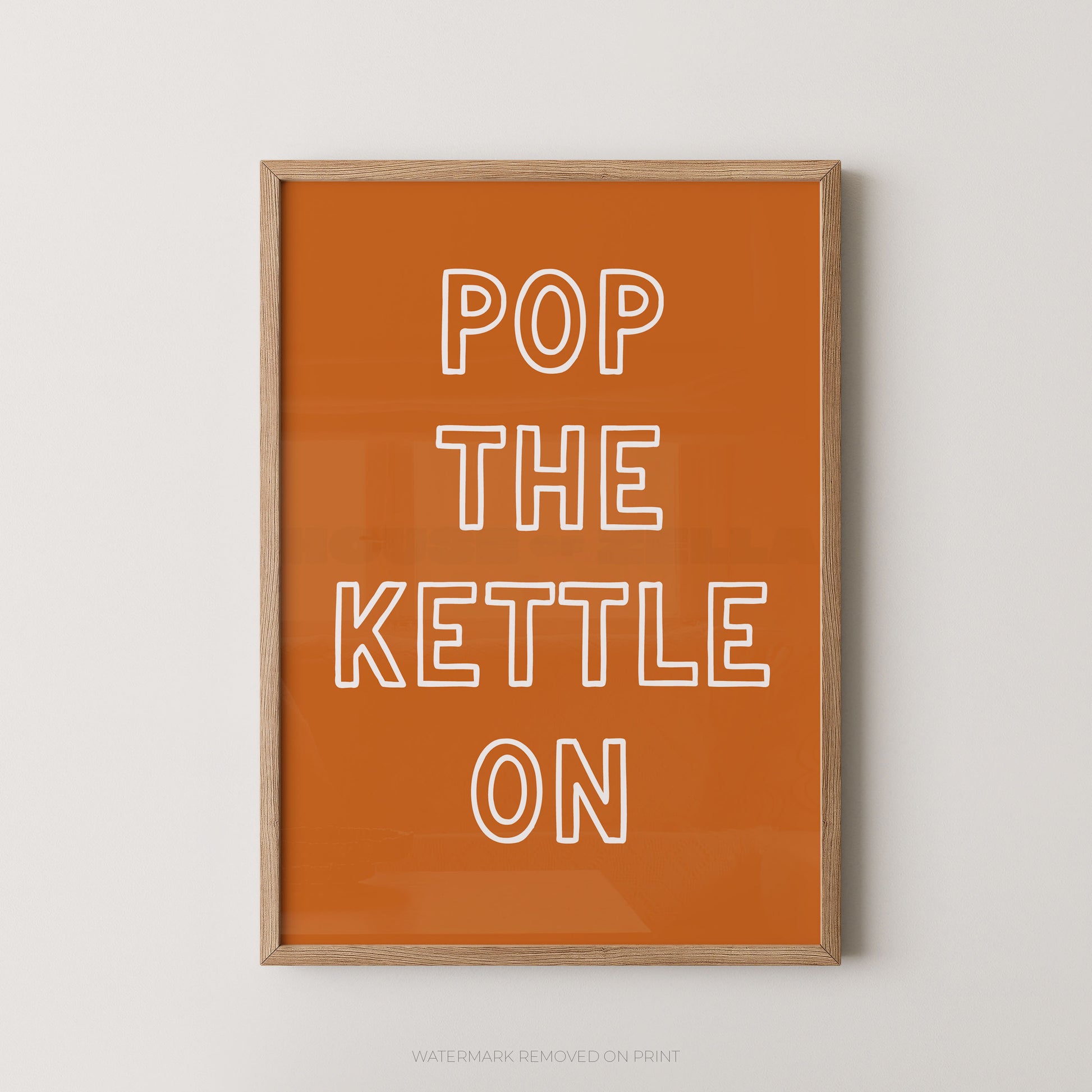 Framed wall art print with the phrase 'POP THE KETTLE ON' in white outlined letters on a warm orange background, shown hanging against a light neutral wall.
