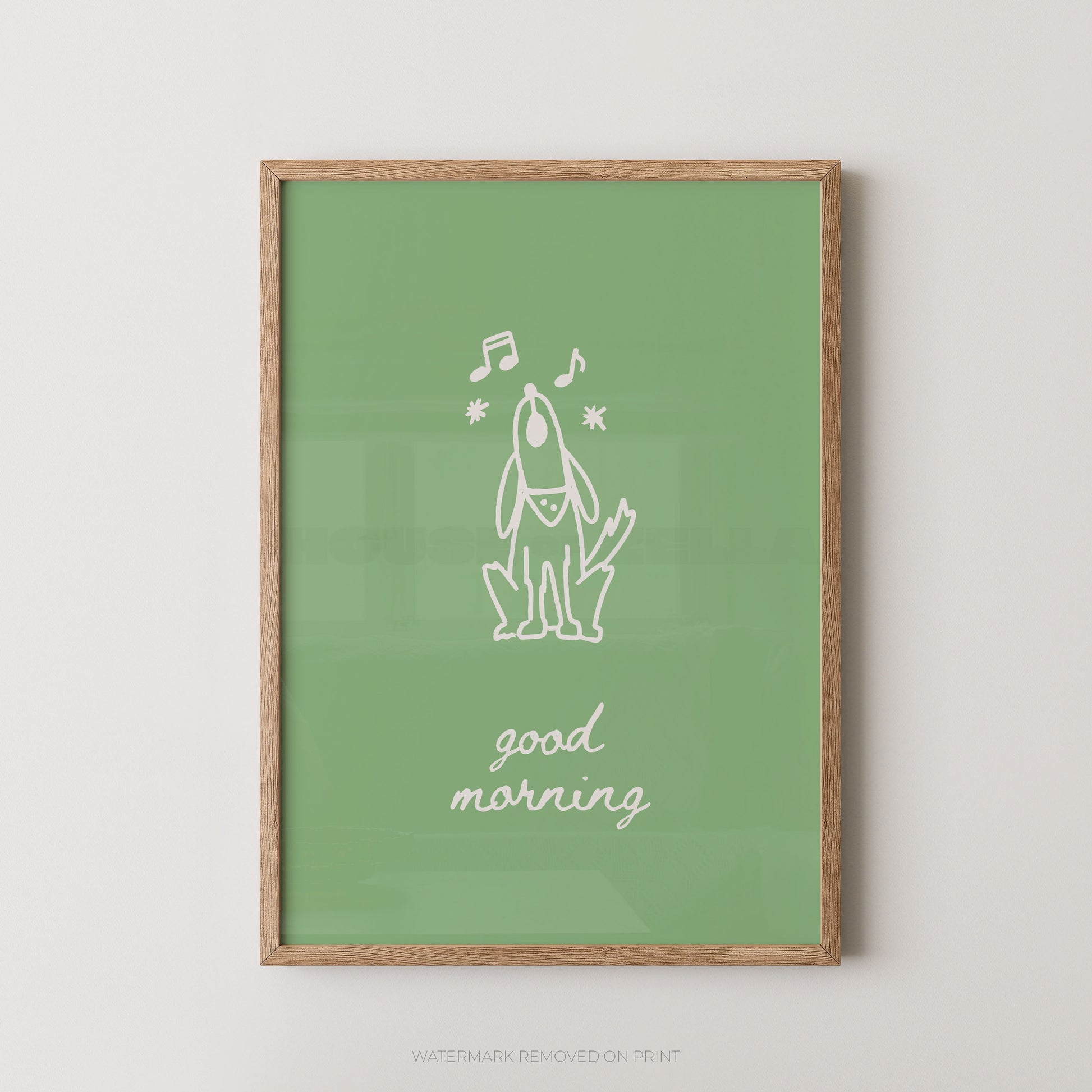A framed art print featuring a minimalist white line drawing of a dog with musical notes and the text 'good morning' on a sage green background, enclosed in a light wood frame against a neutral light gray wall.