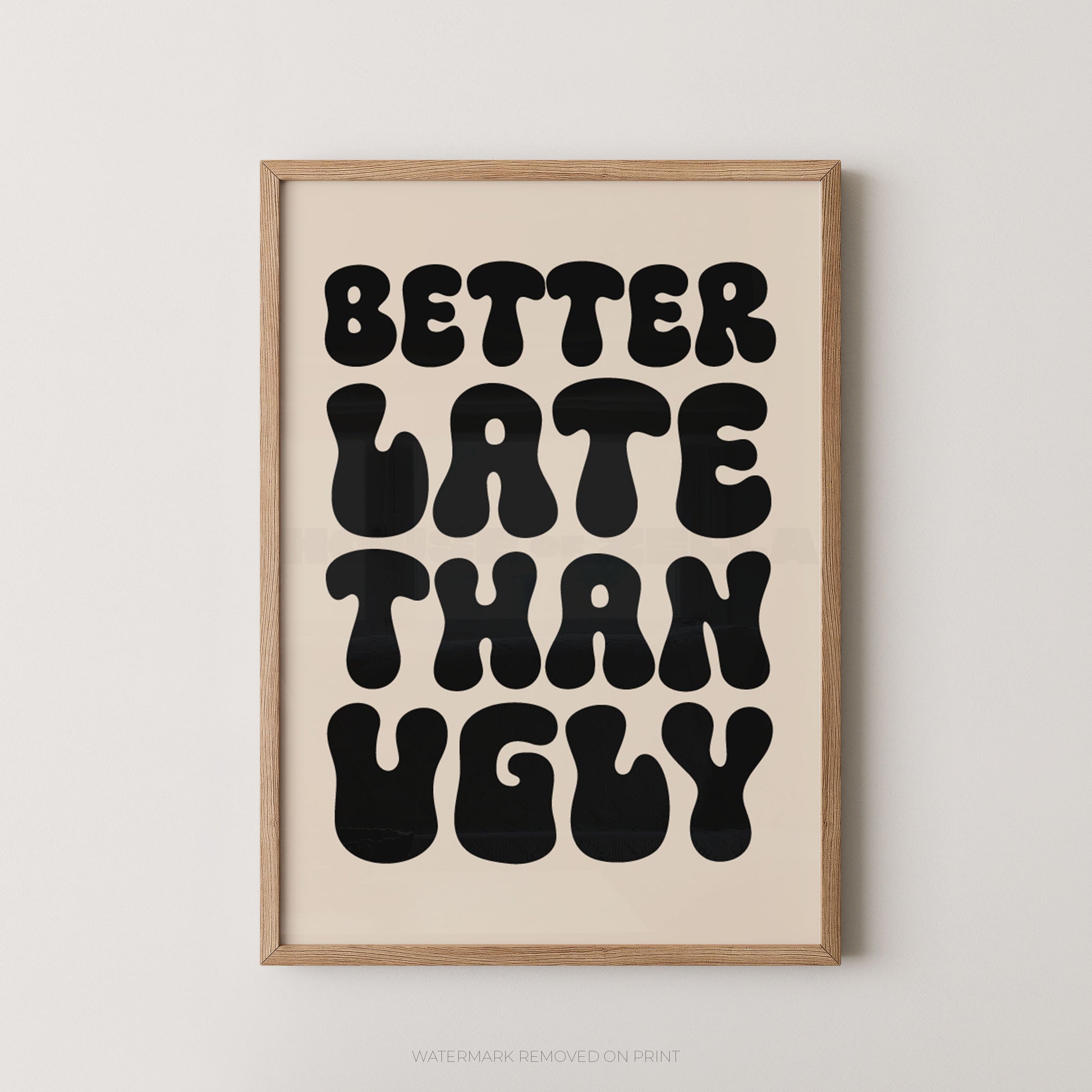 A framed typographic wall art print with the phrase “BETTER LATE THAN UGLY” in large black retro letters on a warm cream background, set in a light natural wood frame against a light gray wall.