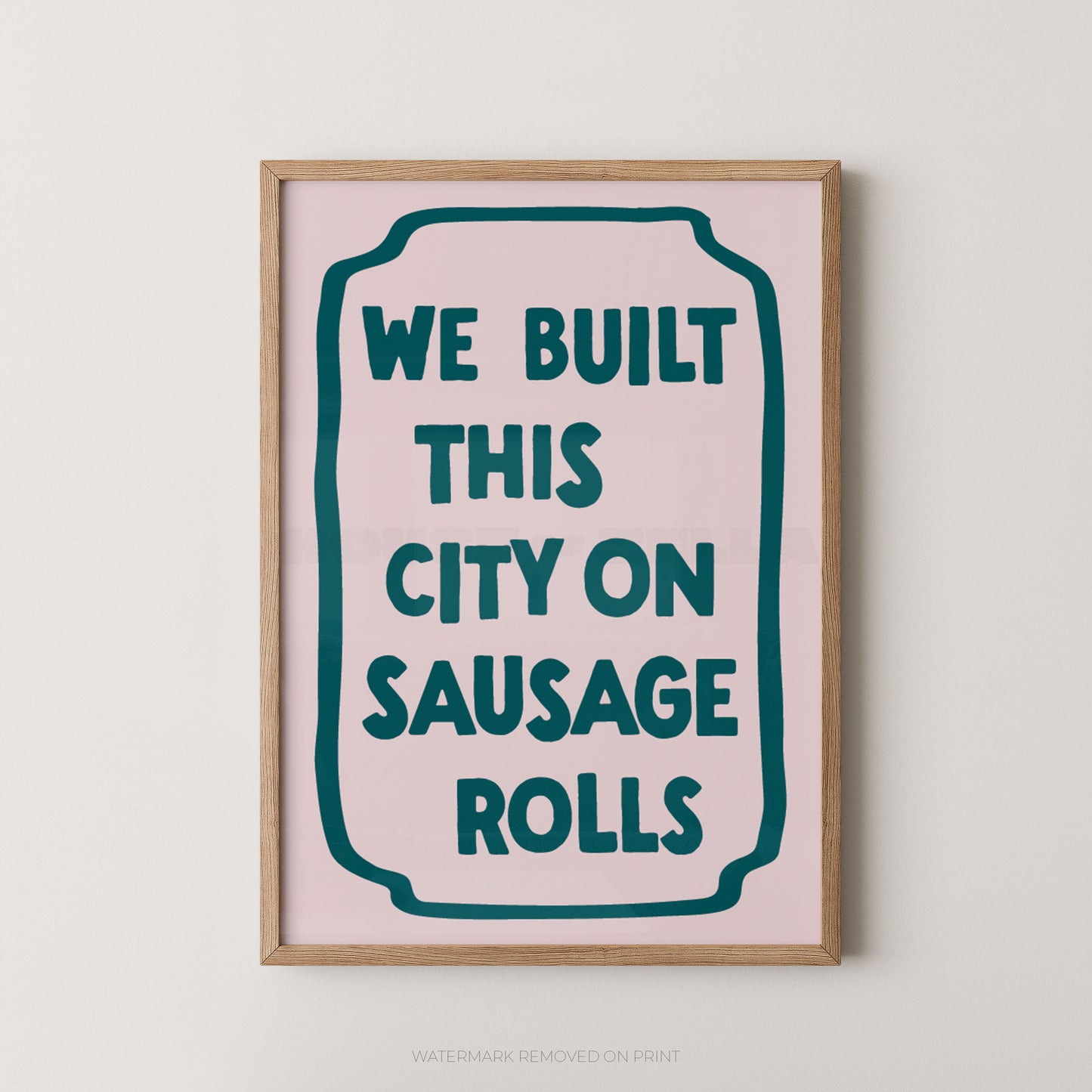A framed typographic wall art print with the phrase “WE BUILT THIS CITY ON SAUSAGE ROLLS” in dark teal lettering on a pale pink background, shown hanging against a light neutral wall.