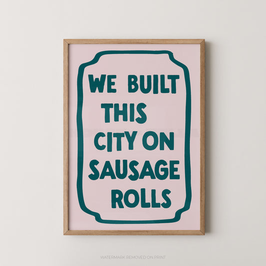 A framed typographic wall art print with the phrase “WE BUILT THIS CITY ON SAUSAGE ROLLS” in dark teal lettering on a pale pink background, shown hanging against a light neutral wall.