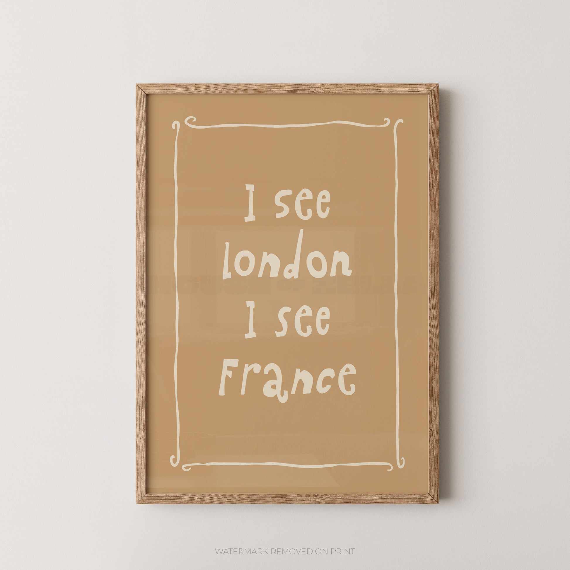 A framed typographic wall art print with the text 'I see London I see France' in white handwritten-style lettering on a warm tan background, enclosed in a light natural wood frame against a light gray wall.
