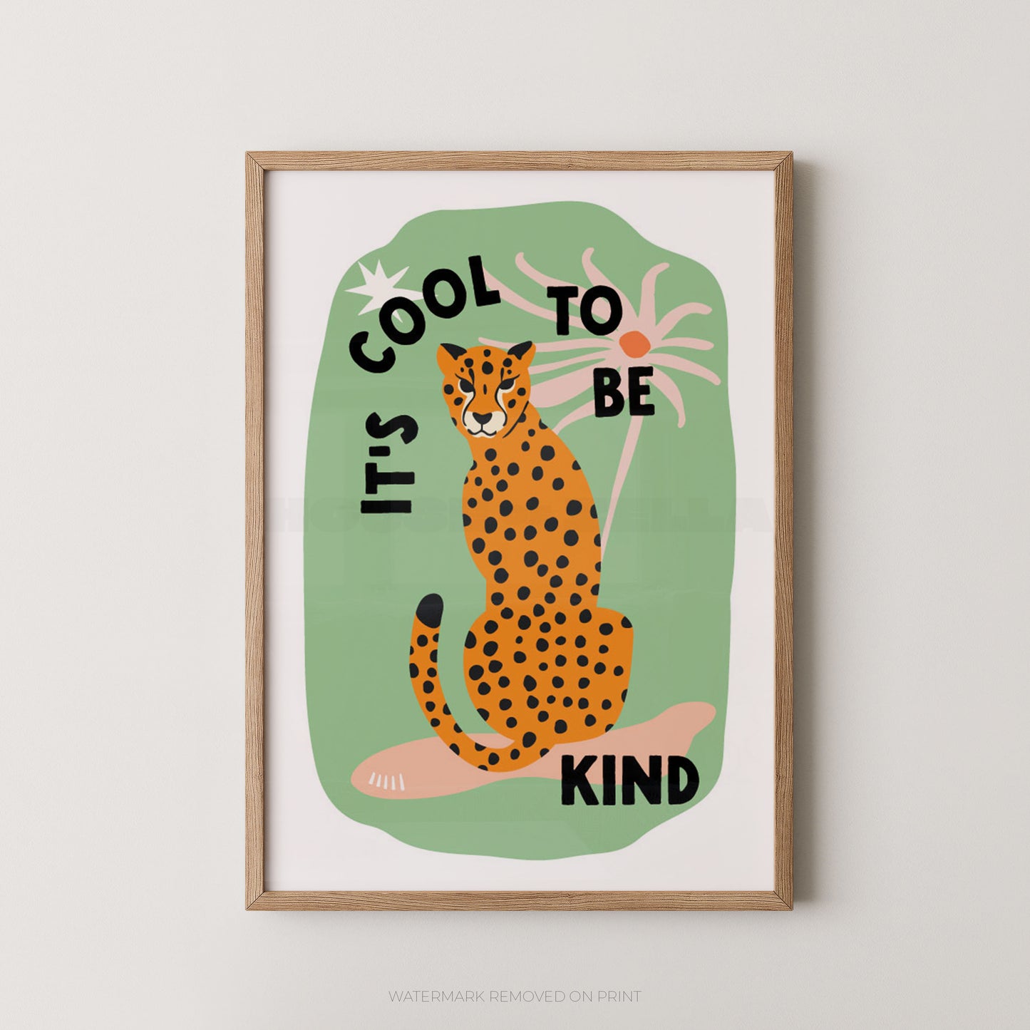 A framed art print featuring a stylized orange and black spotted cheetah sitting on a pink rock against a light green background, with the text 'IT'S COOL TO BE KIND' in black. The print is mounted in a light natural wood frame and hangs on a light gray wall.