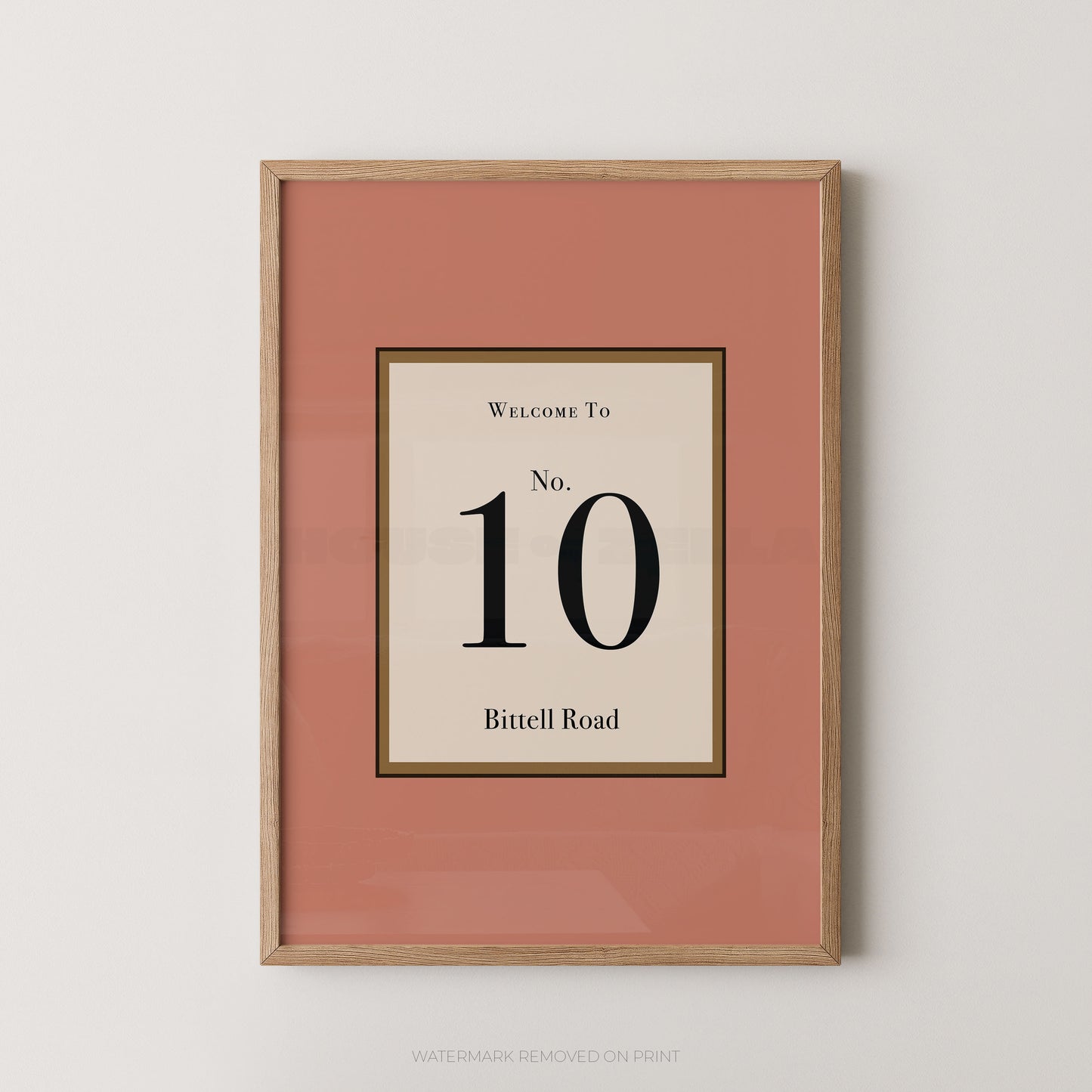 Framed wall art print with a muted pink border and a central cream panel reading 'Welcome To No. 10 Bittell Road' in black text, shown in a light wood frame against a light gray wall.