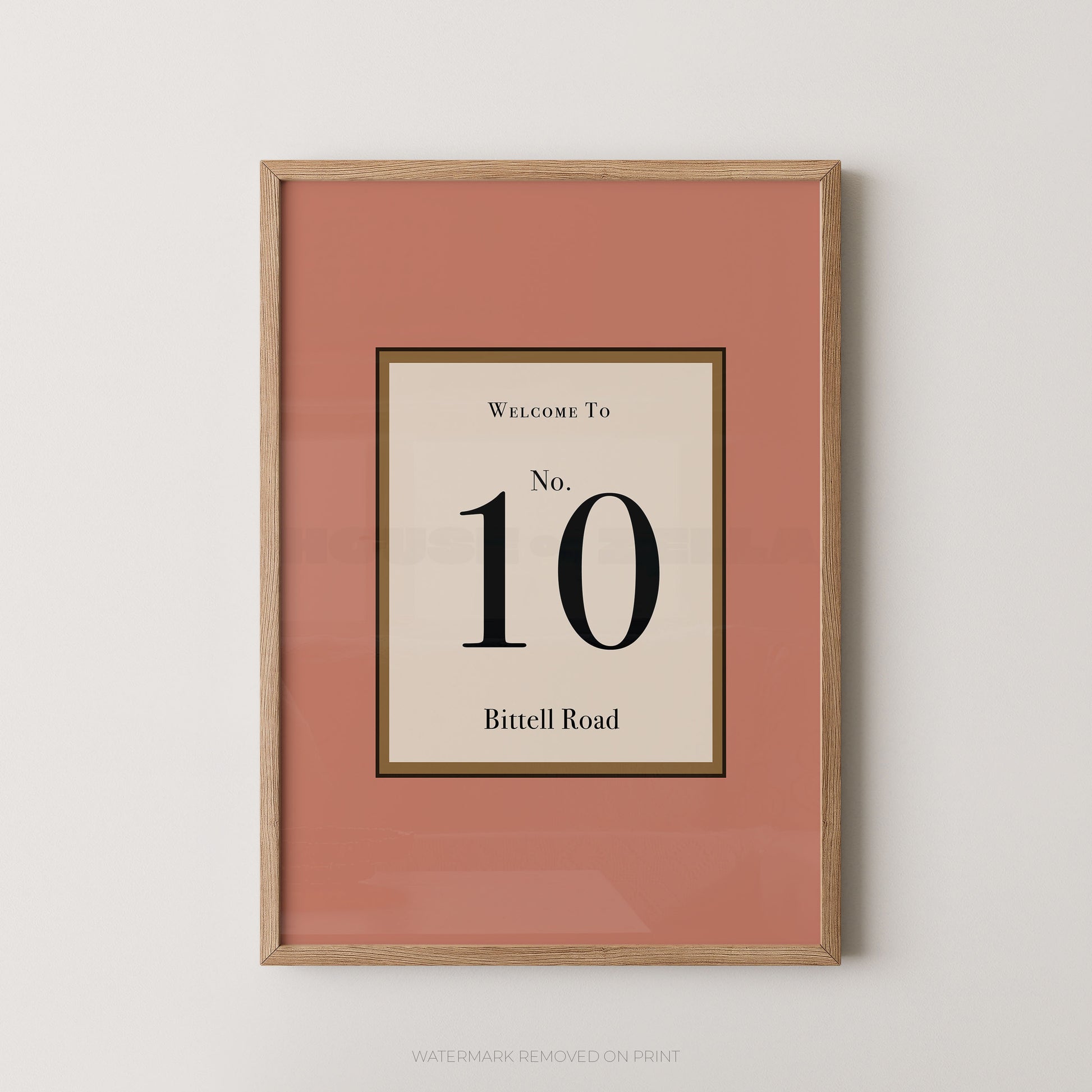 Framed wall art print with a muted pink border and a central cream panel reading 'Welcome To No. 10 Bittell Road' in black text, shown in a light wood frame against a light gray wall.