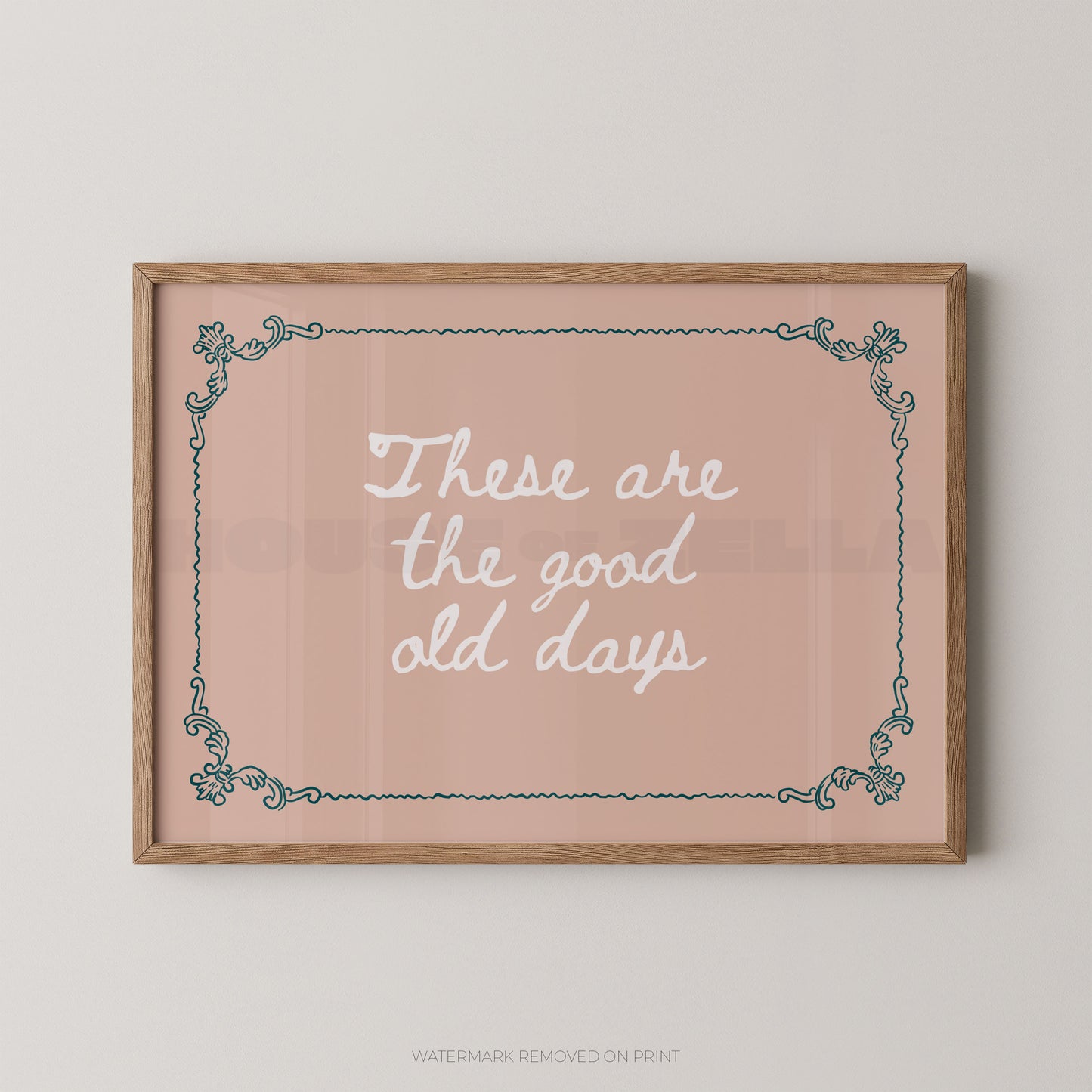 A framed typographic wall art print with the phrase “These are the good old days” in white script on a dusty pink background, bordered by a light wood frame and teal decorative corner flourishes, shown hanging on a light neutral wall.