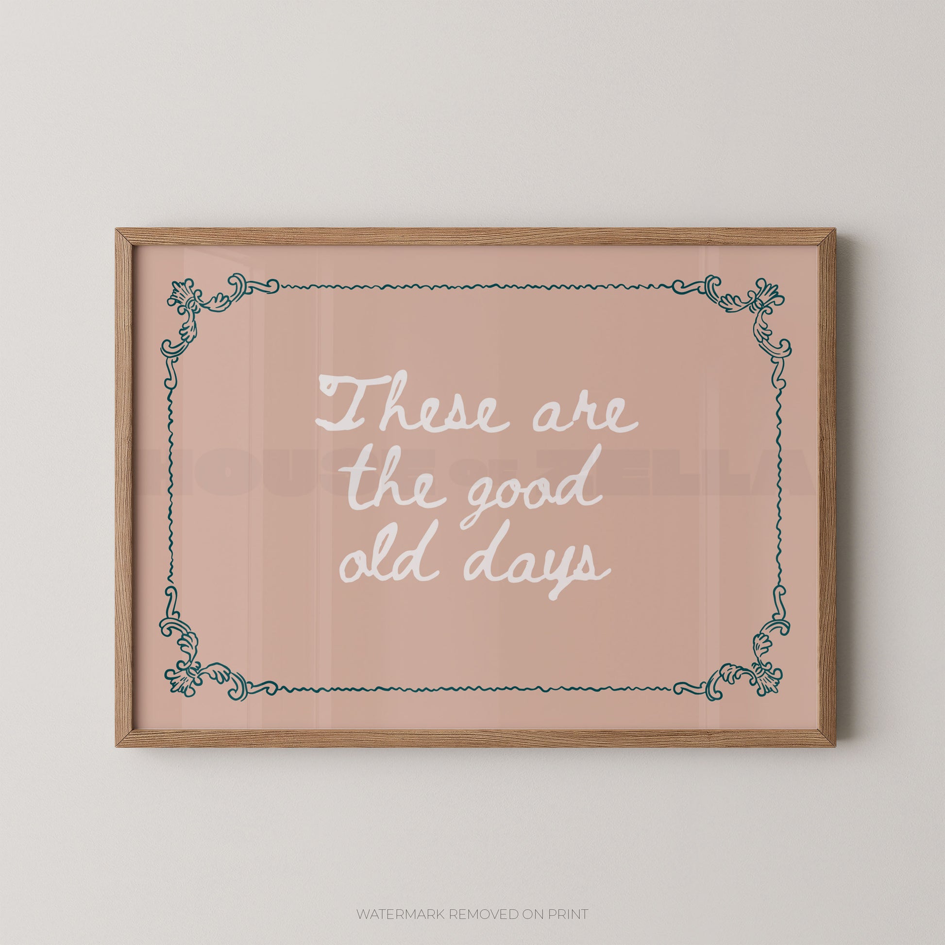 A framed typographic wall art print with the phrase “These are the good old days” in white script on a dusty pink background, bordered by a light wood frame and teal decorative corner flourishes, shown hanging on a light neutral wall.