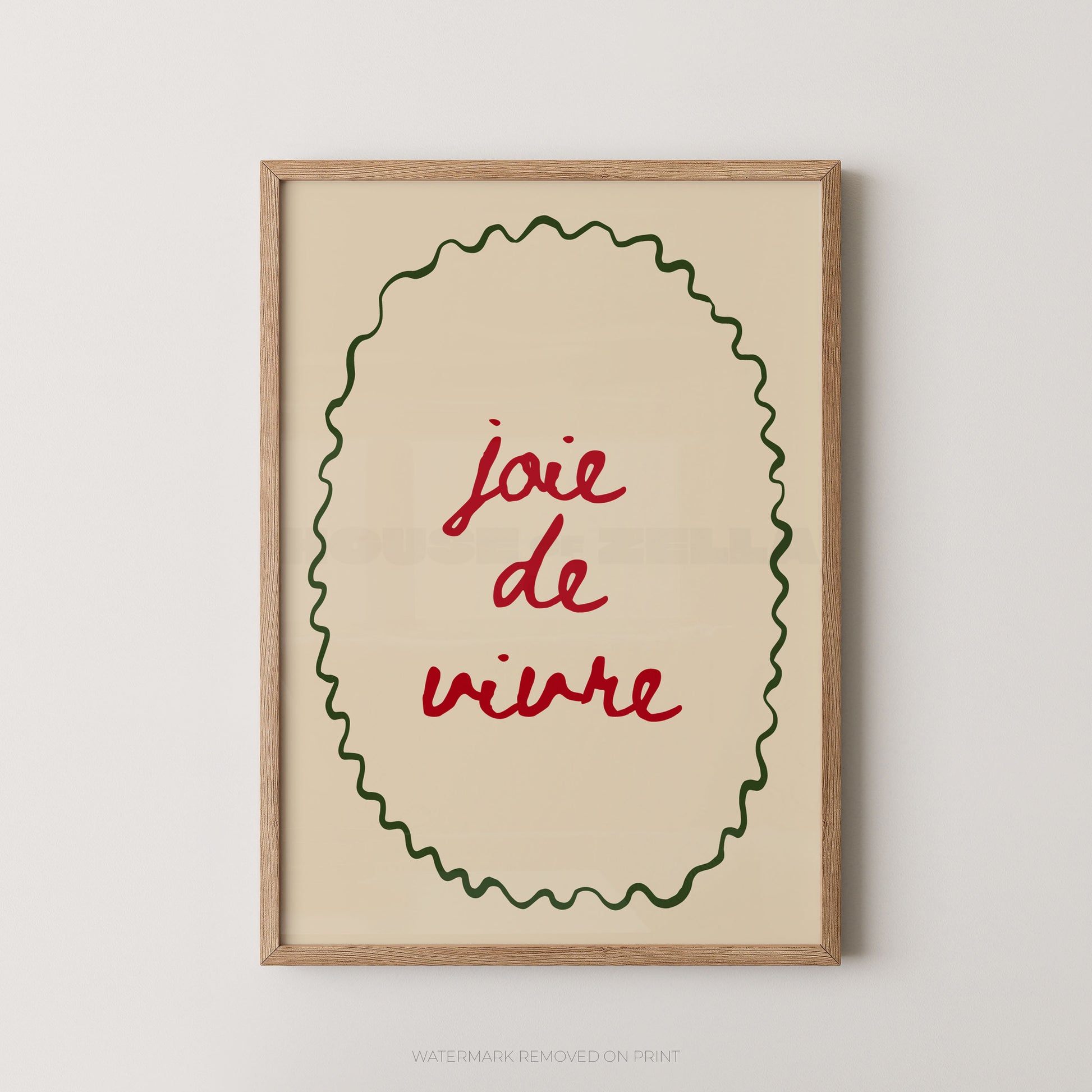 Framed wall art print with the phrase “joie de vivre” in red script inside a green wavy oval border on a warm cream background, shown in a light wood frame against a light gray wall.