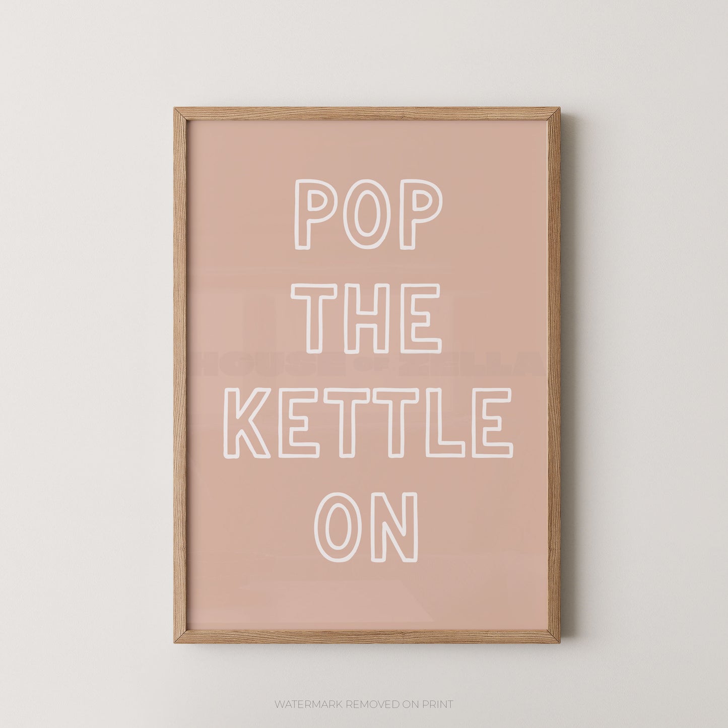 A framed typographic wall art print with the phrase “POP THE KETTLE ON” in white outlined letters on a dusty pink background, shown in a light natural wood frame against a light gray wall.