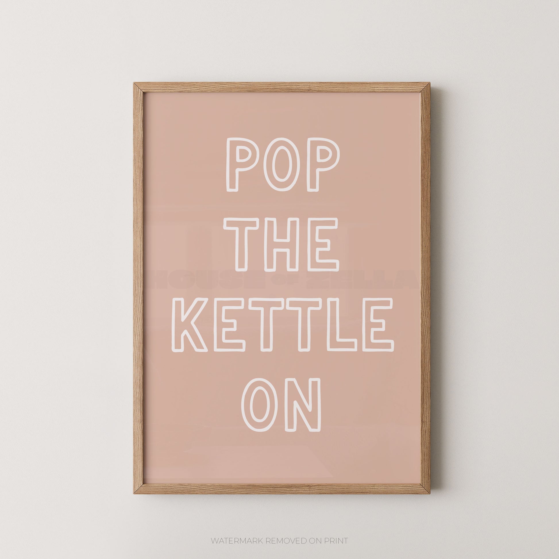 A framed typographic wall art print with the phrase “POP THE KETTLE ON” in white outlined letters on a dusty pink background, shown in a light natural wood frame against a light gray wall.
