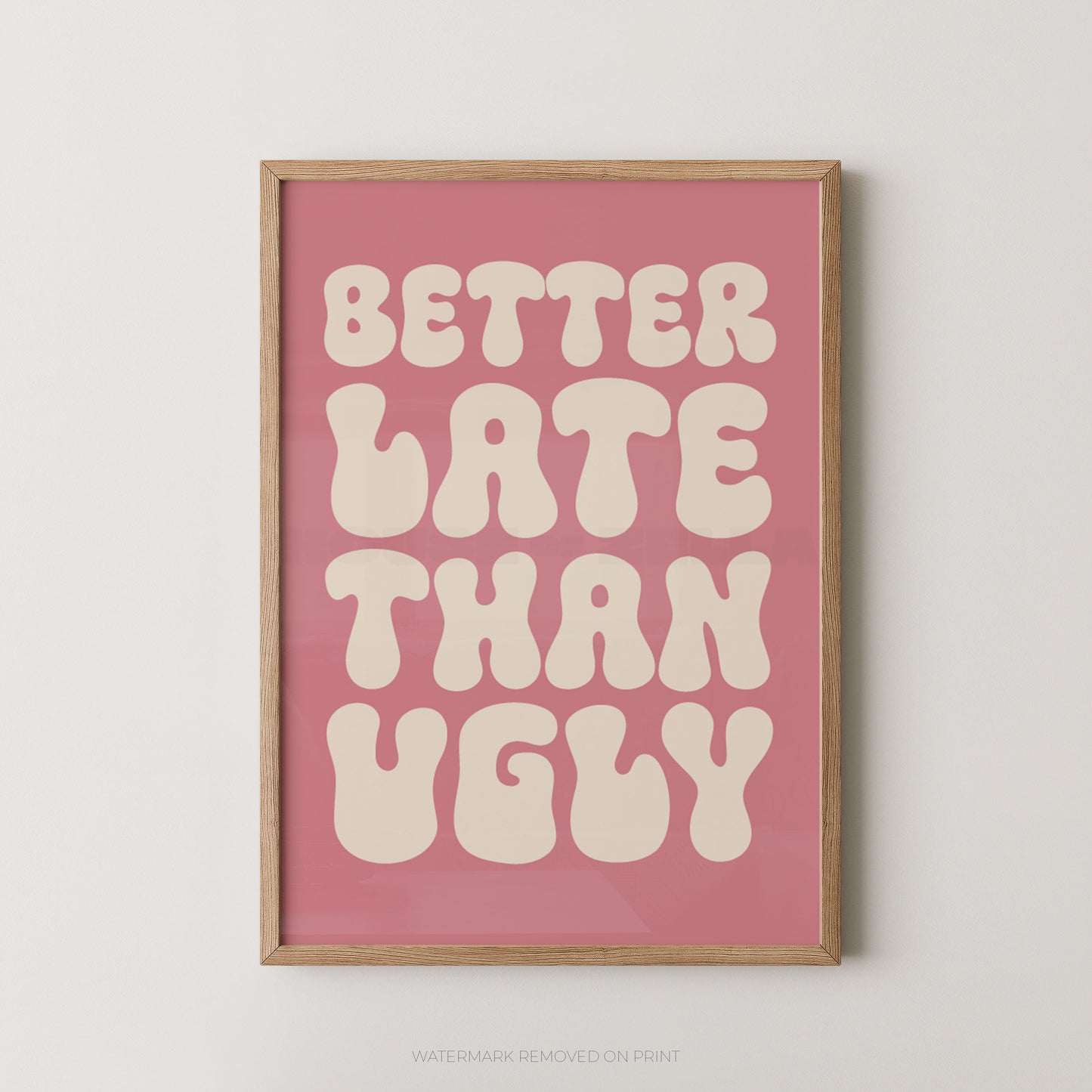 A framed typographic wall art print with the phrase “BETTER LATE THAN UGLY” in large cream bubble letters on a dusty pink background, shown hanging against a light neutral wall.