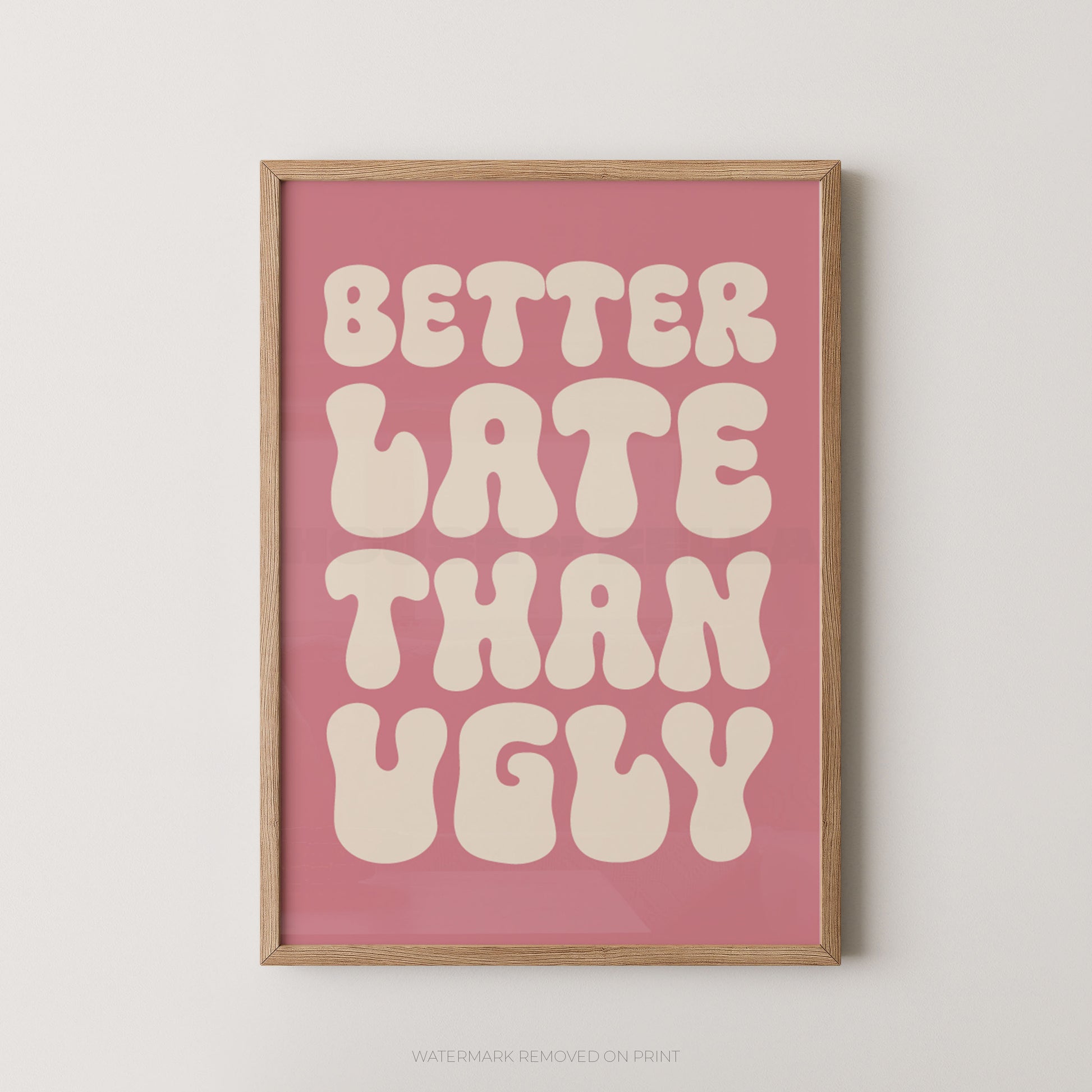 A framed typographic wall art print with the phrase “BETTER LATE THAN UGLY” in large cream bubble letters on a dusty pink background, shown hanging against a light neutral wall.