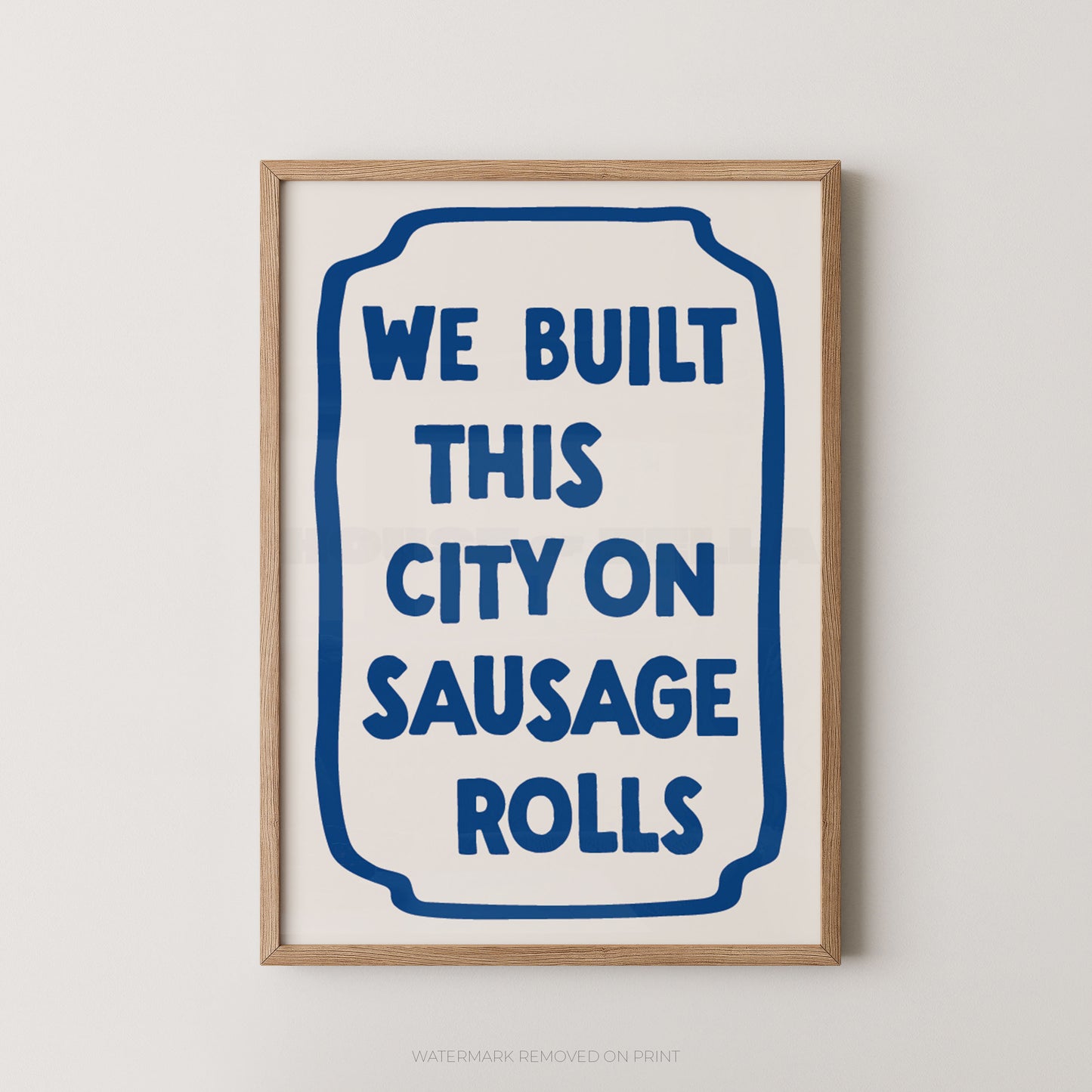 A framed typographic wall art print with the phrase “WE BUILT THIS CITY ON SAUSAGE ROLLS” in blue text inside a rounded blue border, shown against a light neutral wall.