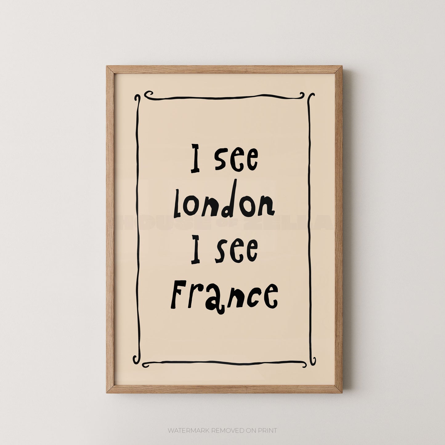Framed typographic wall art with the text 'I see London I see France' in black handwritten-style lettering on a warm cream background, enclosed in a light natural wood frame against a light gray wall.