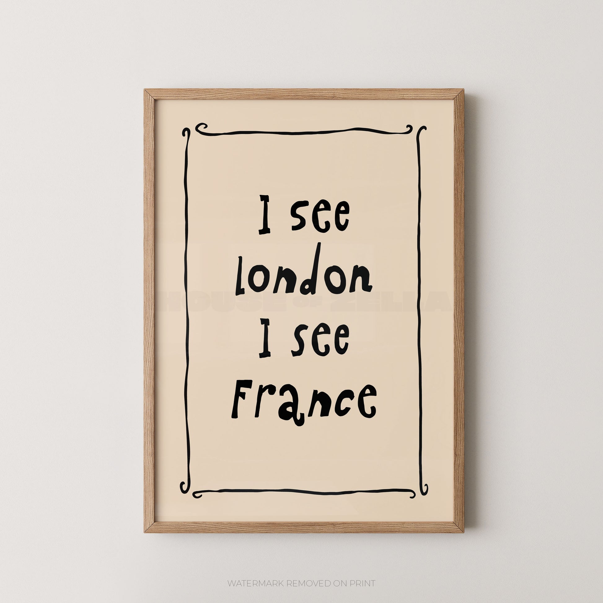Framed typographic wall art with the text 'I see London I see France' in black handwritten-style lettering on a warm cream background, enclosed in a light natural wood frame against a light gray wall.