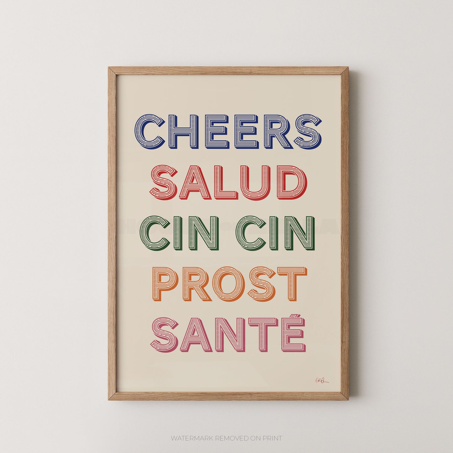A framed typographic wall art print with the words 'CHEERS SALUD CIN CIN PROST SANTÉ' in multicolored letters on a cream background, shown hanging against a light neutral wall.