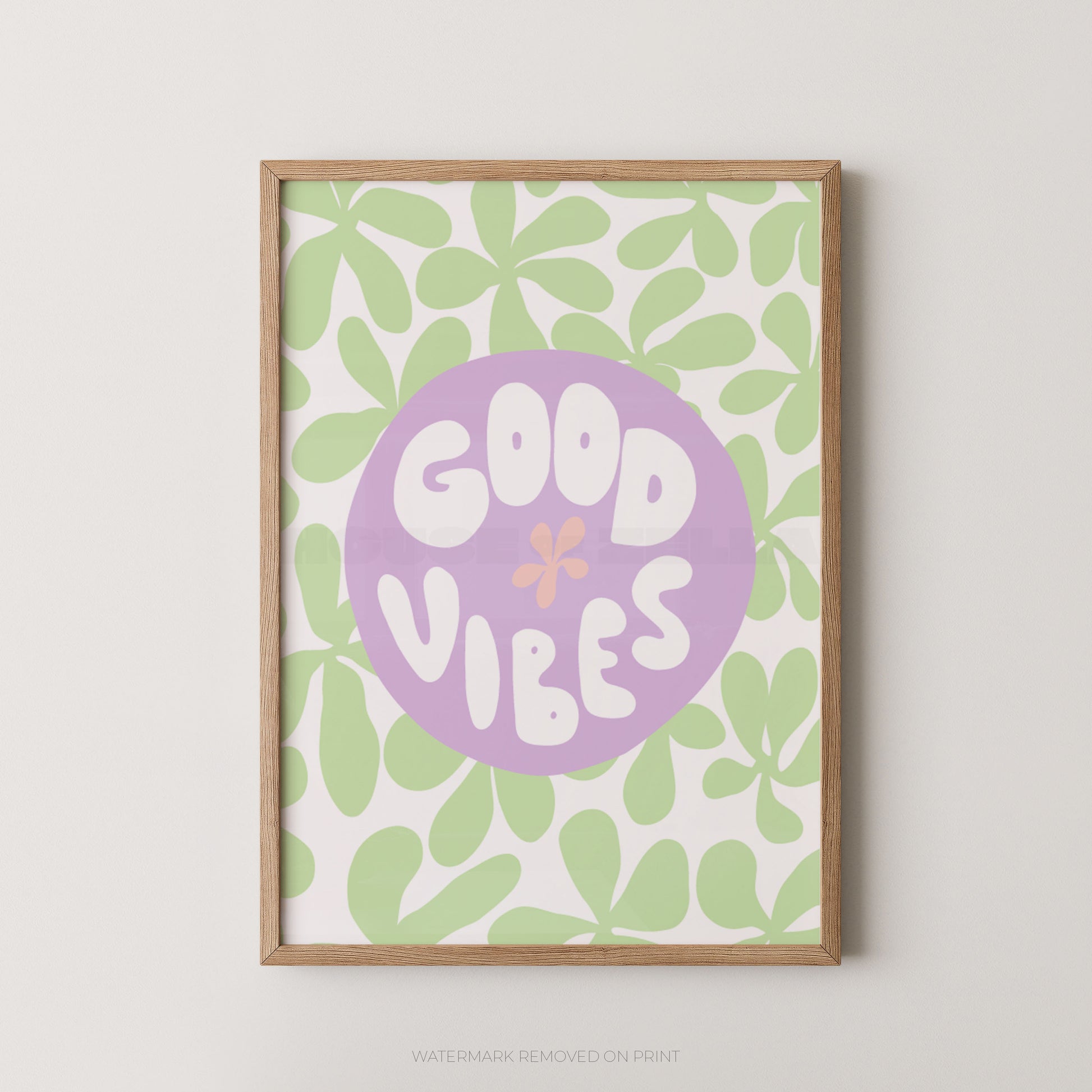 A framed art print featuring a retro pattern of light green leaves on a white background, with a central lavender purple circle containing the words 'GOOD VIBES' in white retro lettering and a small pink flower accent. The print is housed in a light natural wood frame and is displayed against a neutral light gray wall.