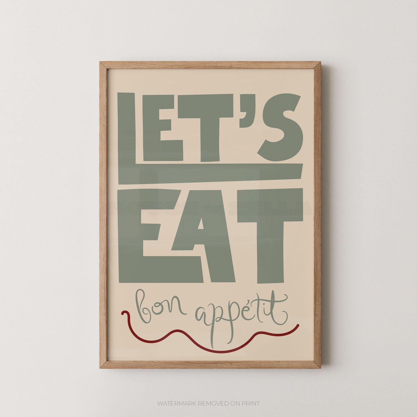 A framed typographic wall art print with the words “LET’S EAT bon appetit” in muted green, cream, and red on a light wood frame, shown against a plain light gray wall.