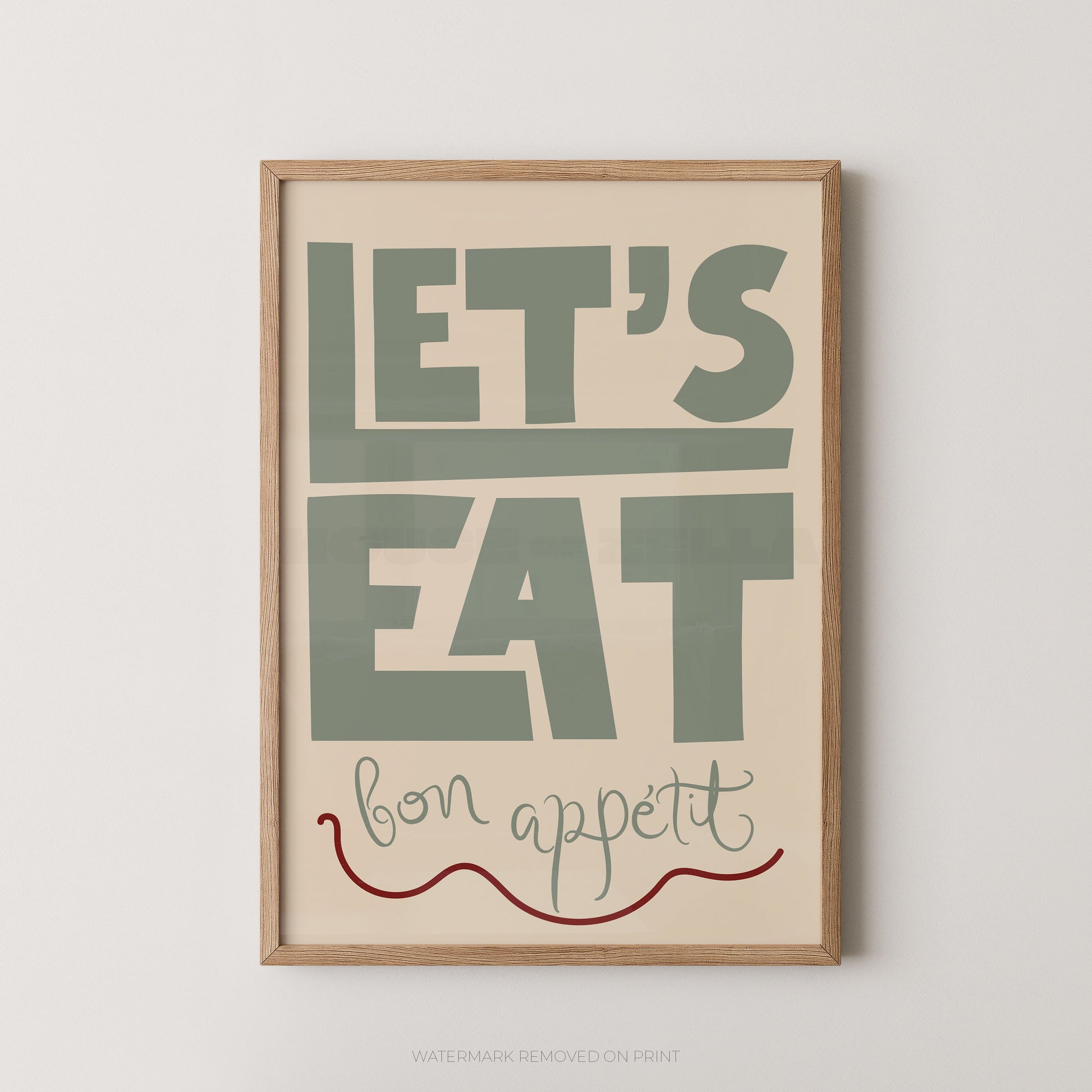A framed typographic wall art print with the words “LET’S EAT bon appetit” in muted green, cream, and red on a light wood frame, shown against a plain light gray wall.