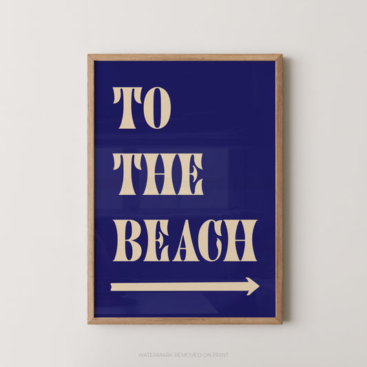 A framed typographic wall art print with the words “TO THE BEACH” in large cream letters on a deep blue background, plus a cream arrow pointing right. The print is shown hanging against a light neutral wall.