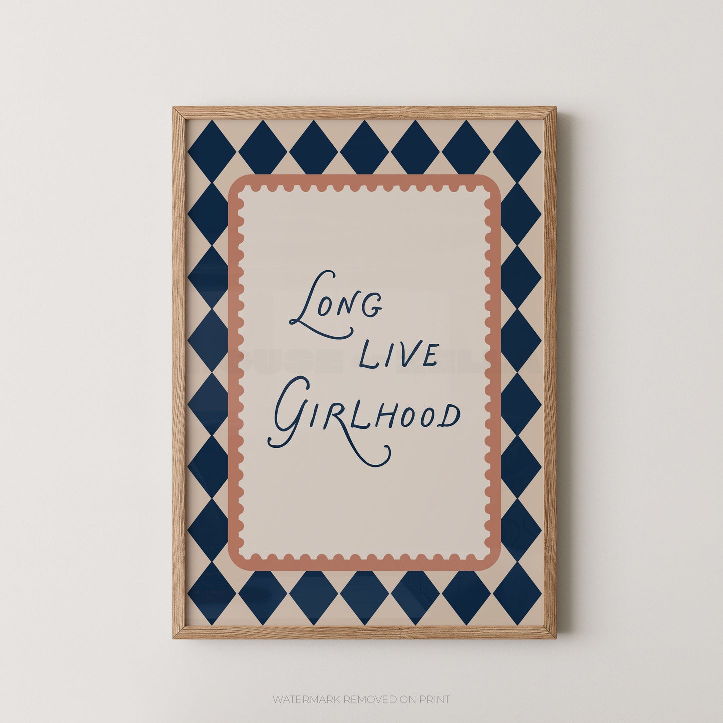 A framed typographic wall art print with the phrase “Long Live Girlhood” in dark blue script, surrounded by a peach scalloped inner border and a navy-and-beige diamond patterned outer border, shown against a light neutral wall.