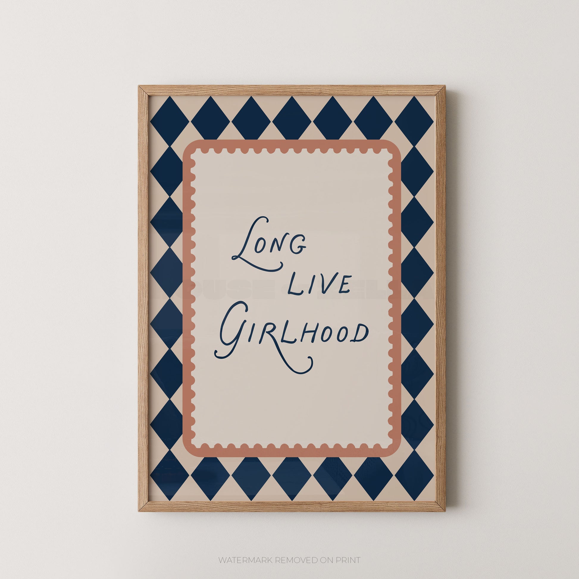 A framed typographic wall art print with the phrase “Long Live Girlhood” in dark blue script, surrounded by a peach scalloped inner border and a navy-and-beige diamond patterned outer border, shown against a light neutral wall.