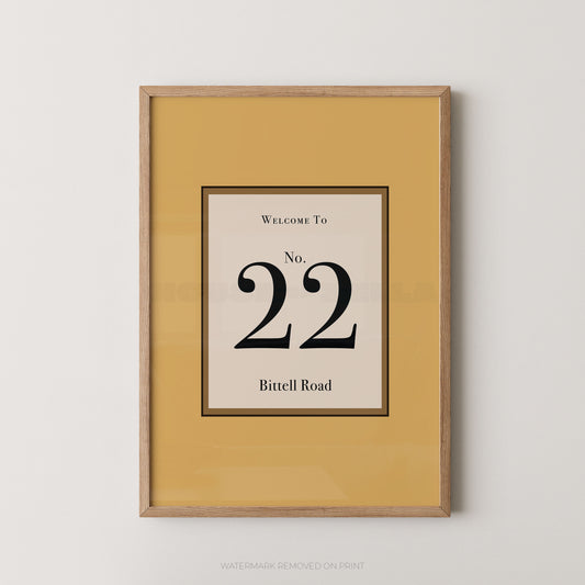 Framed wall art print with a mustard yellow border and a central cream panel reading 'Welcome To No. 22 Bittell Road' in black text, shown in a light wood frame against a light gray wall.