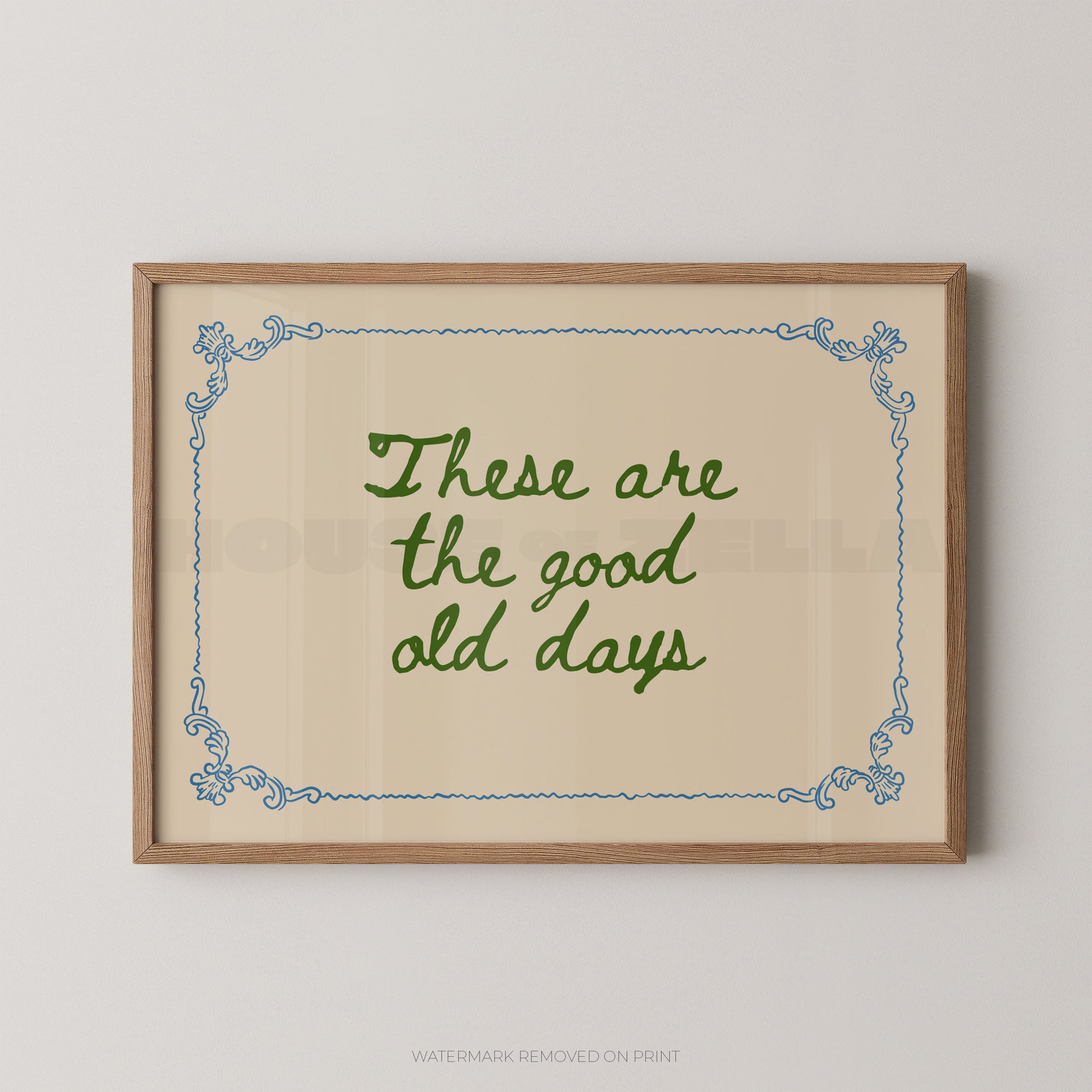 Framed wall art print with the phrase “These are the good old days” in green script on a beige background, bordered by a light wood frame and blue decorative corner flourishes, shown hanging on a light gray wall.