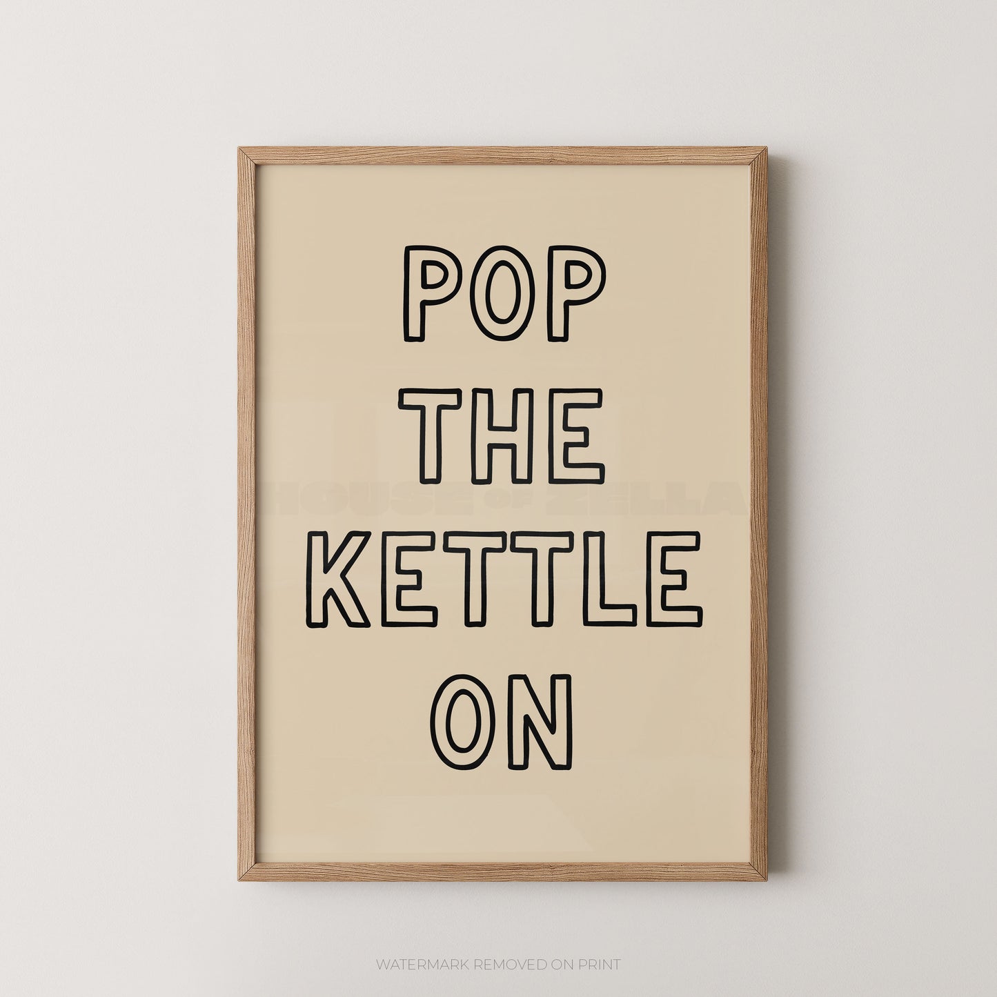 A framed typographic wall art print with the phrase “POP THE KETTLE ON” in black outlined letters on a warm cream background, shown in a light natural wood frame against a light gray wall.