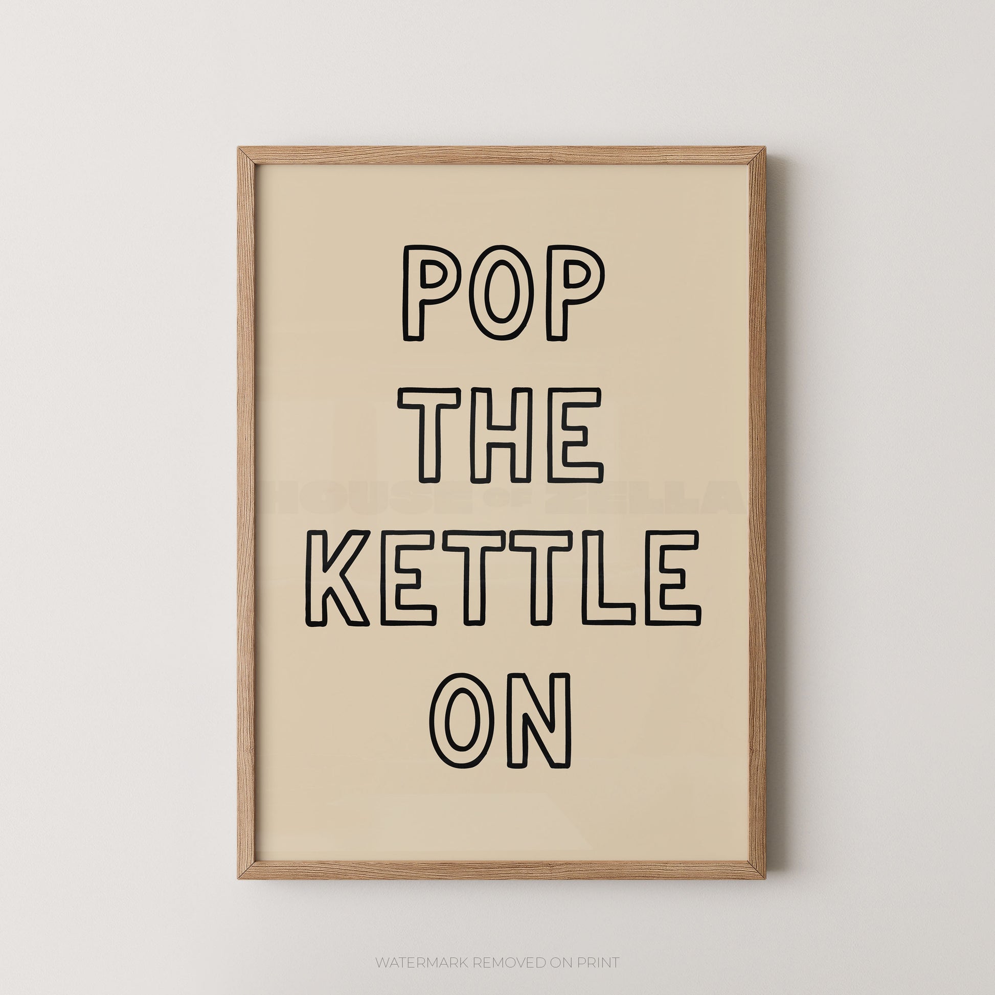 A framed typographic wall art print with the phrase “POP THE KETTLE ON” in black outlined letters on a warm cream background, shown in a light natural wood frame against a light gray wall.