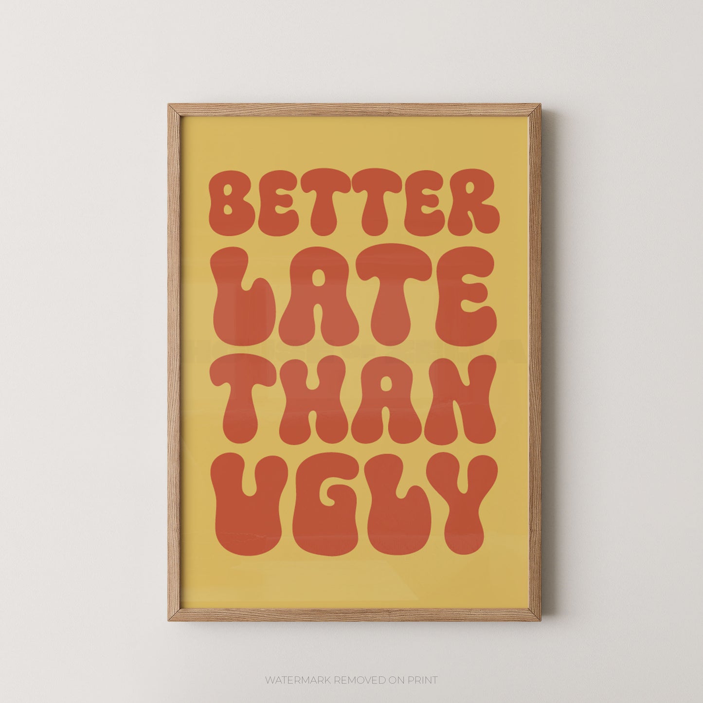A framed typographic wall art print with the phrase “BETTER LATE THAN UGLY” in large retro red-orange letters on a warm yellow background, shown hanging against a light neutral wall.