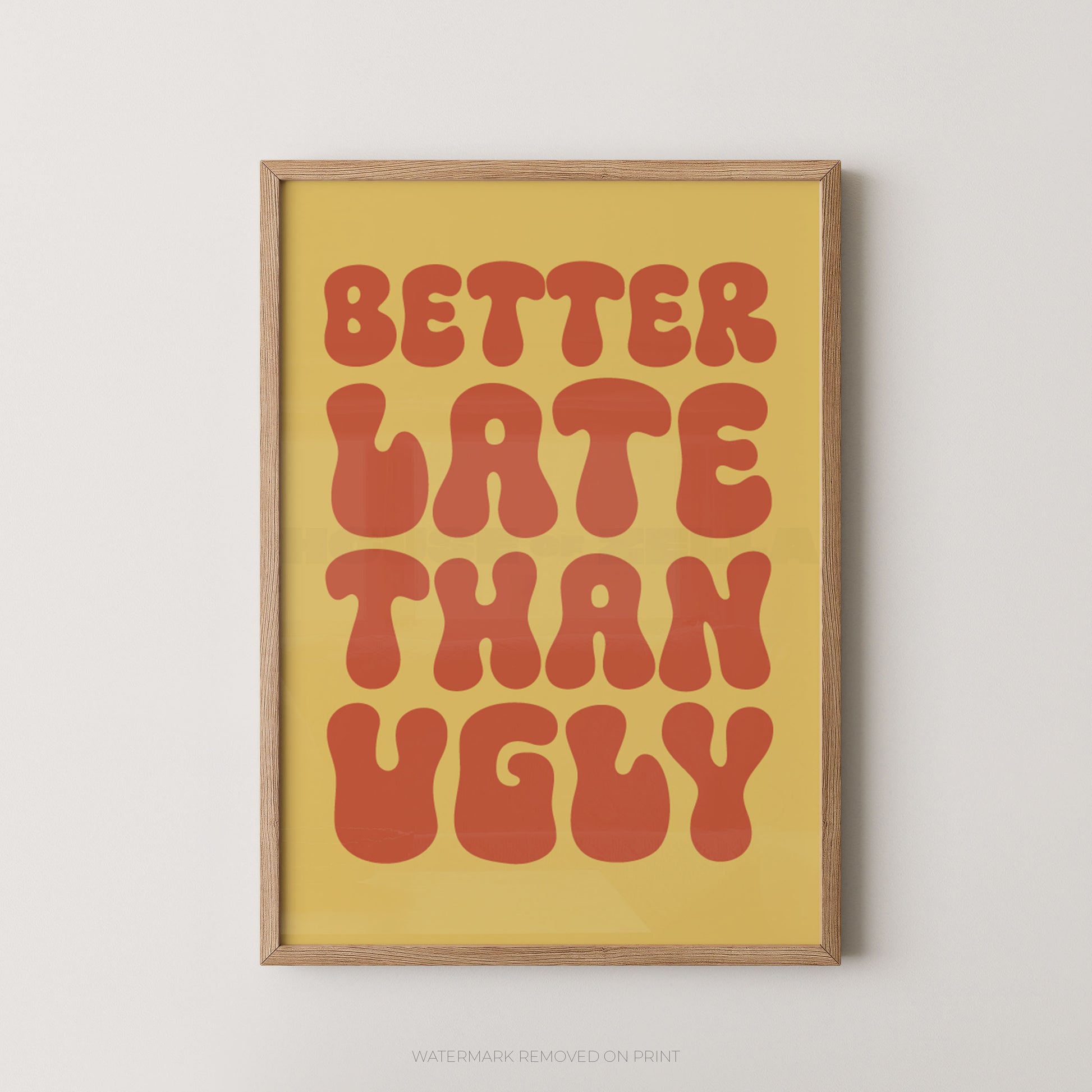 A framed typographic wall art print with the phrase “BETTER LATE THAN UGLY” in large retro red-orange letters on a warm yellow background, shown hanging against a light neutral wall.