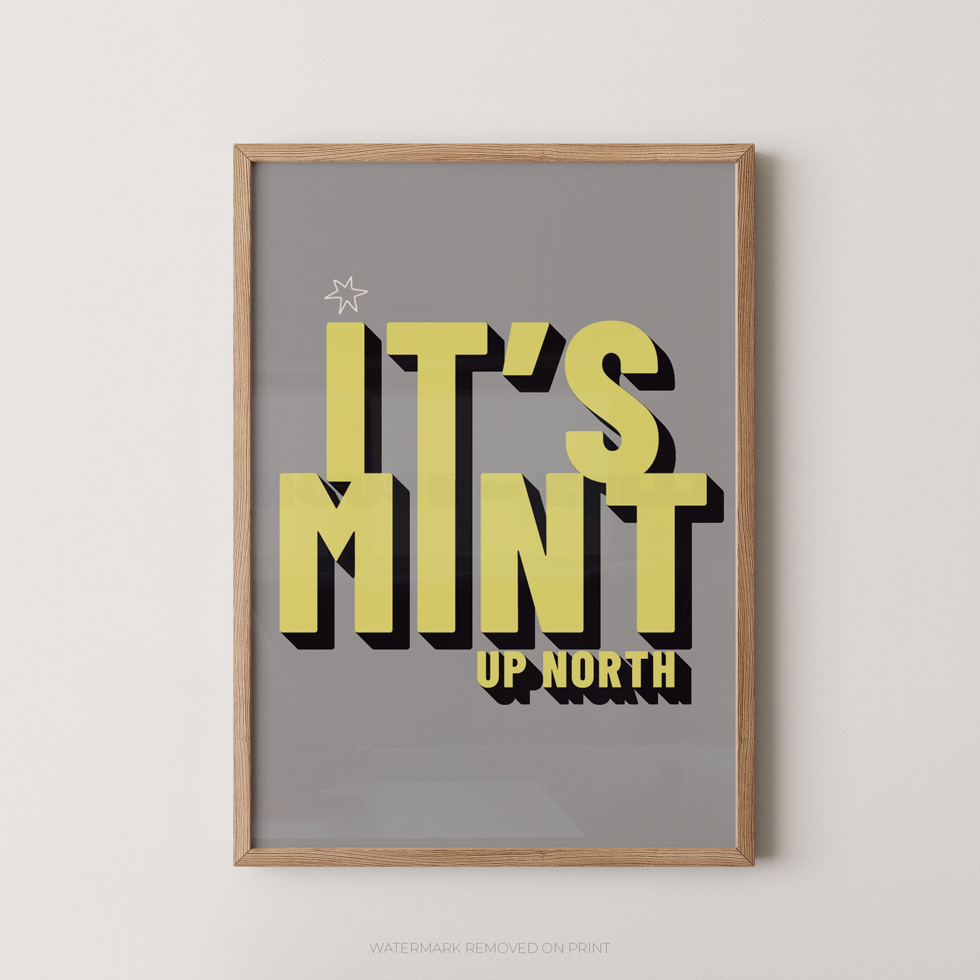 Framed typographic wall art reading “IT’S MINT UP NORTH” in large yellow letters with black drop shadows on a gray background, shown in a light wood frame against a light neutral wall.
