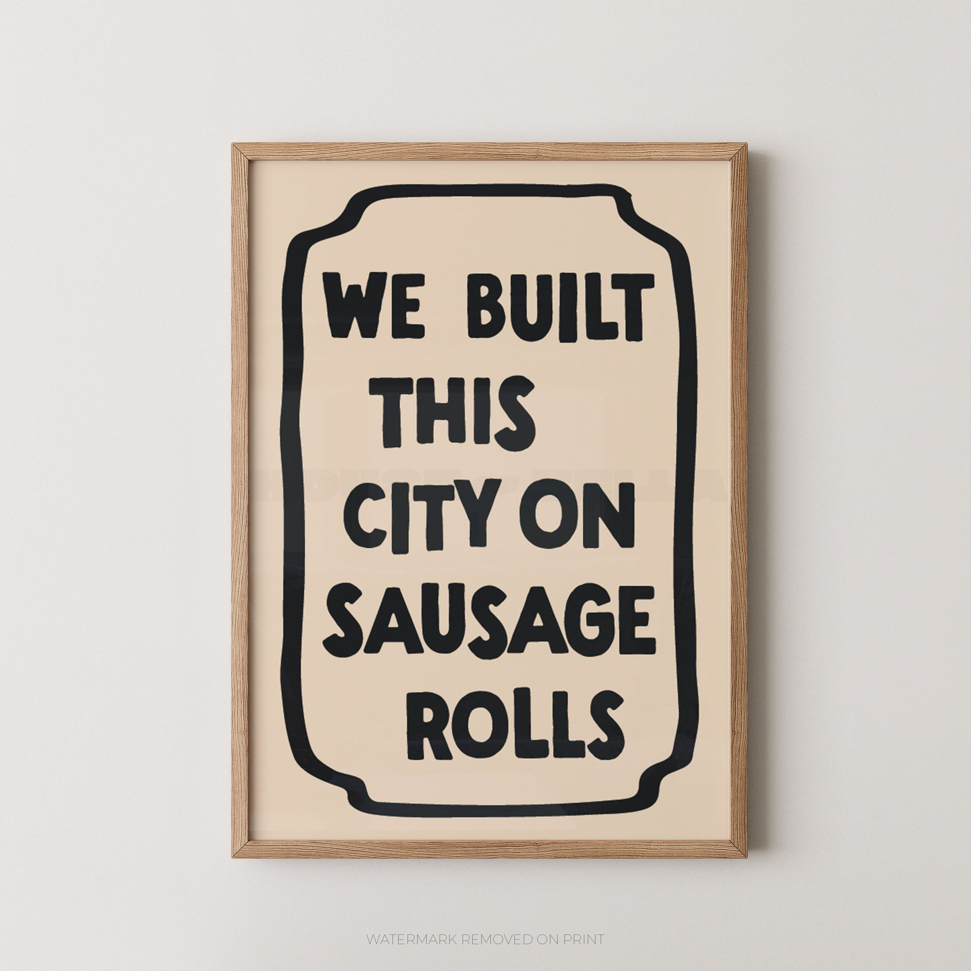 Framed wall art print with the phrase “WE BUILT THIS CITY ON SAUSAGE ROLLS” in bold black text inside a cream rectangle, set in a light natural wood frame against a light gray wall.
